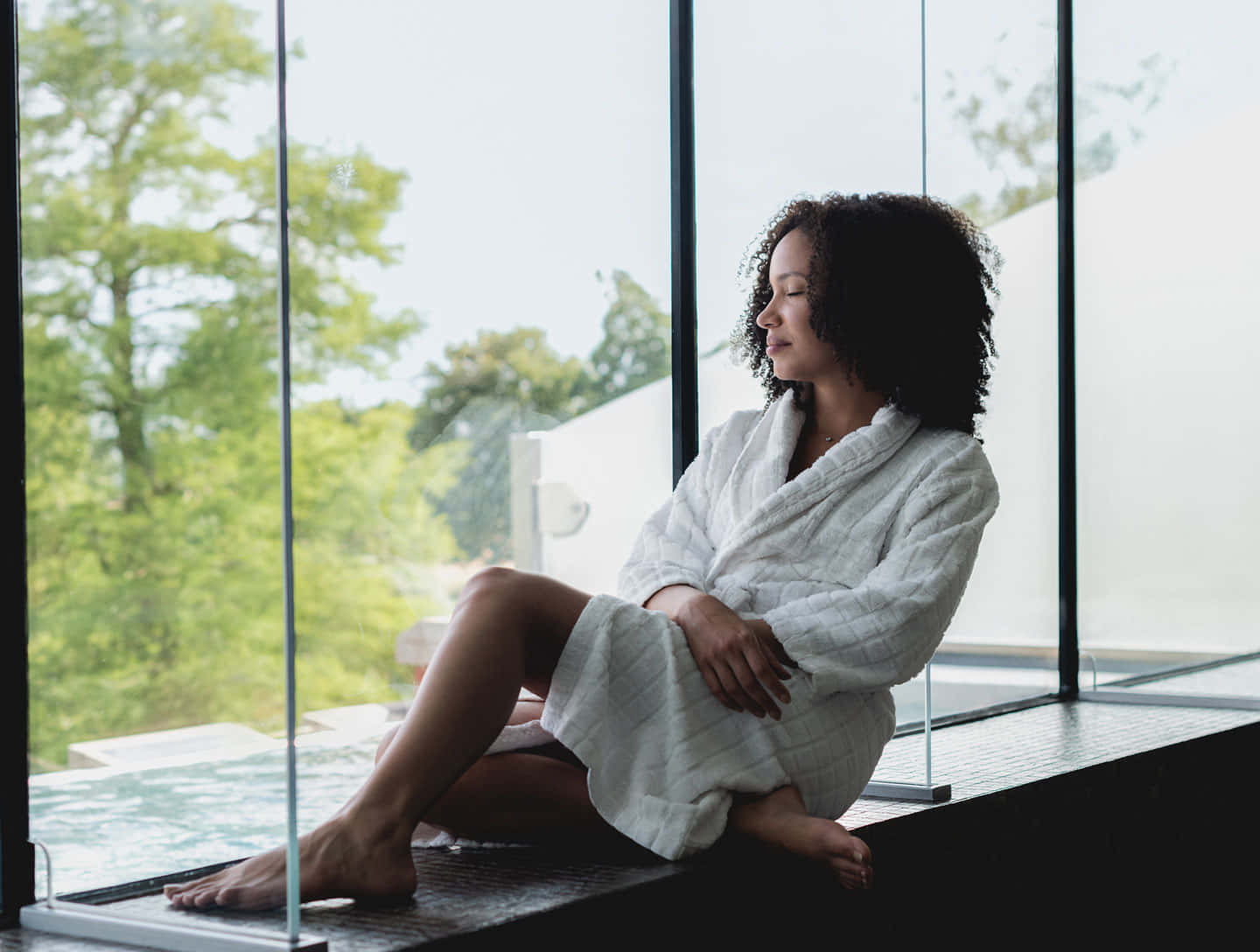 Woman 桌布 壁紙 — 一位身穿白色長袍的女性赤腳坐在玻璃邊緣，平靜地凝視著外面的綠樹。