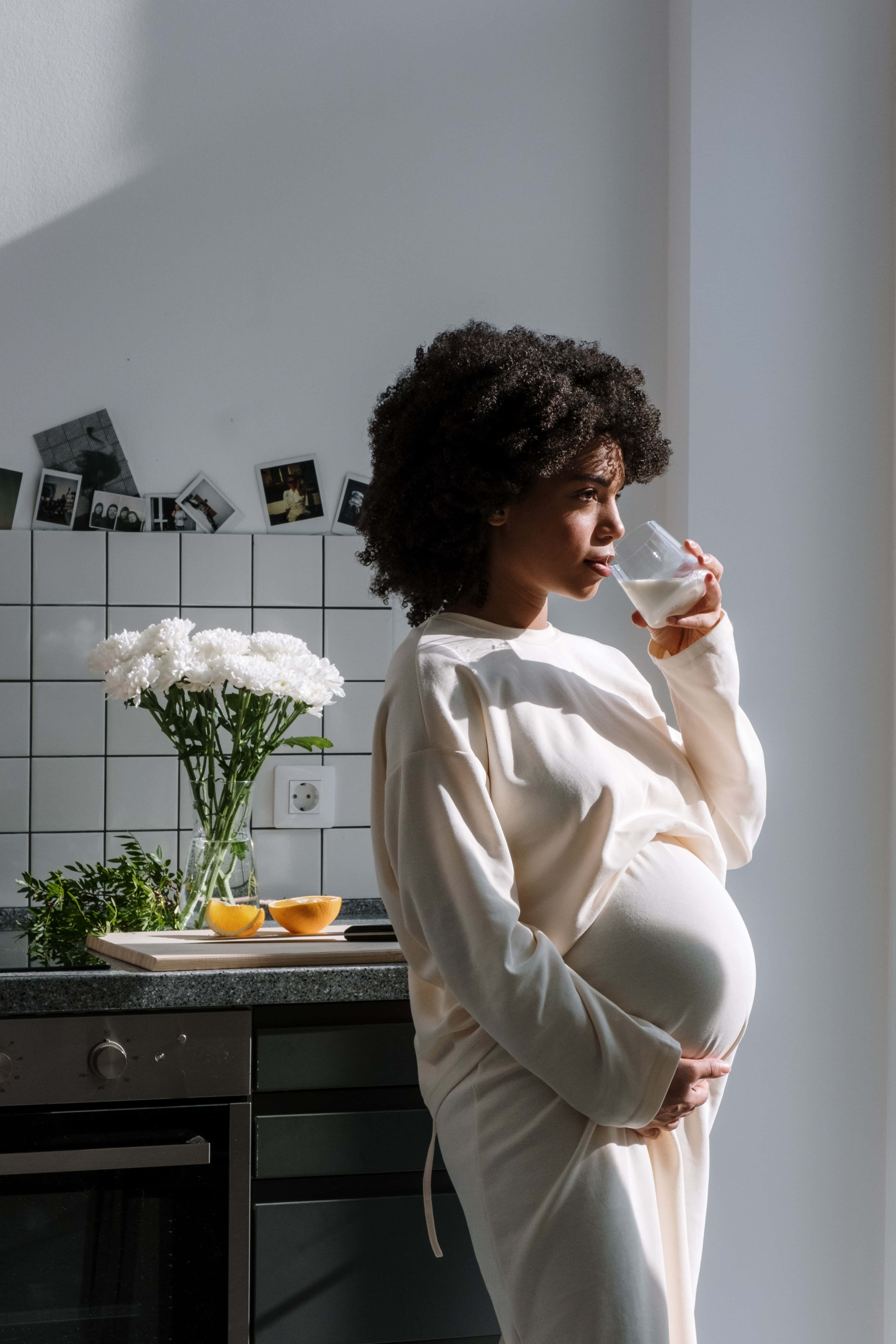 Pregnant 桌布 壁紙 — 一位身穿白色長袍的孕婦在陽光明媚的廚房裡，旁邊放著一瓶白花，小口喝著牛奶。