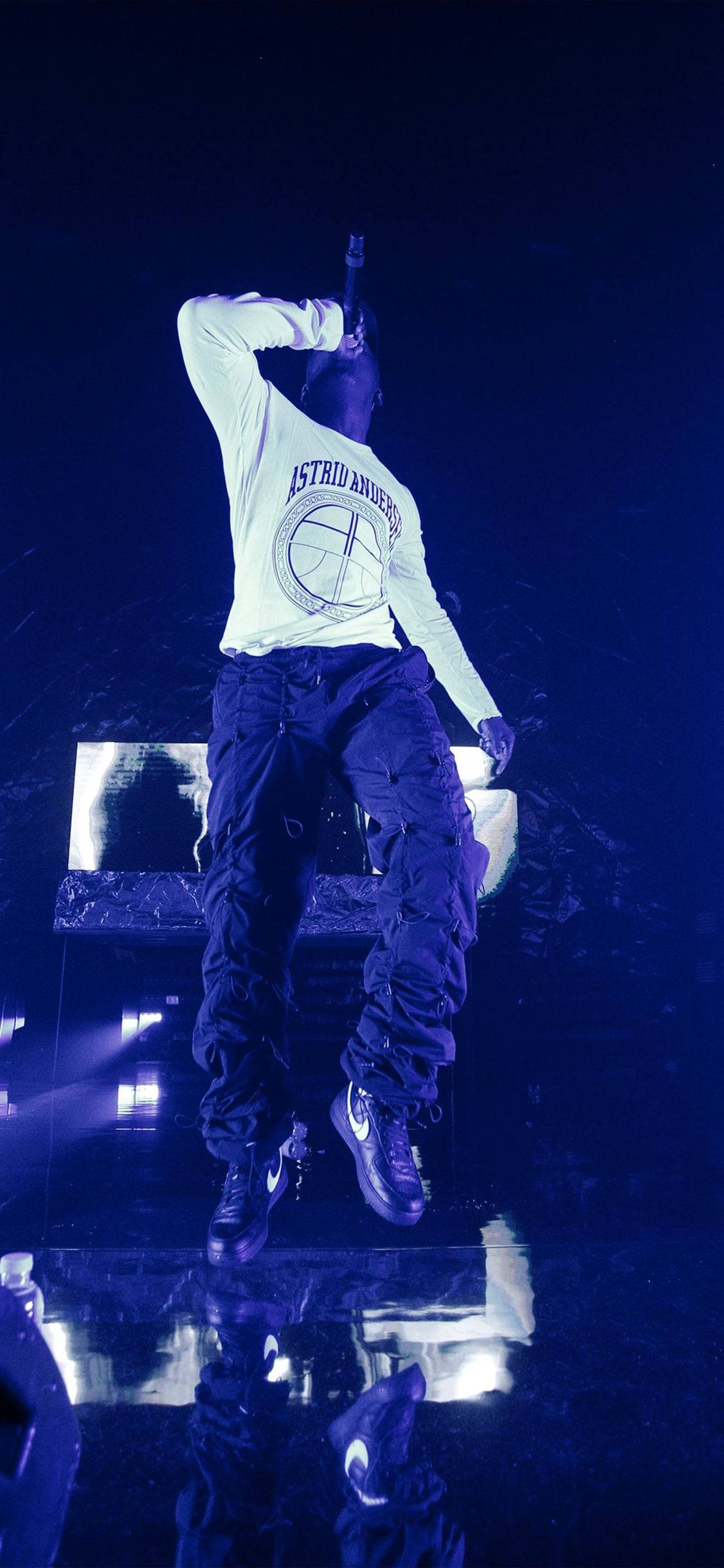 Skepta 桌布 壁紙 — 一位身穿白色長袖上衣和深色褲子的表演者站在反射舞台上，在強烈的藍色燈光下舉起一隻手臂。