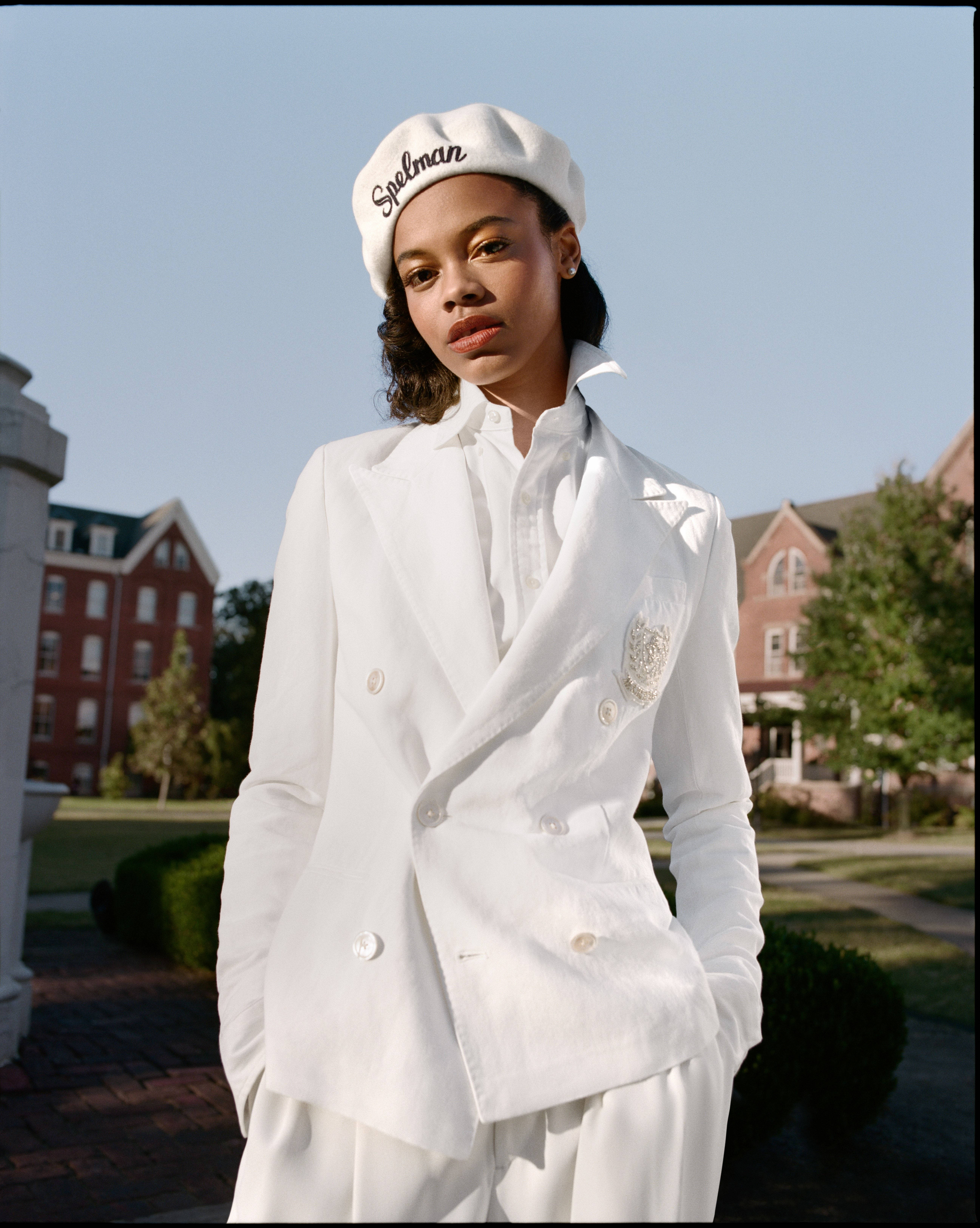 白色 桌布 壁紙 — 一位身穿白色雙排扣西裝和「Spelman」貝雷帽的優雅女士，在晴朗藍天下自信地站在磚砌建築和綠植前。