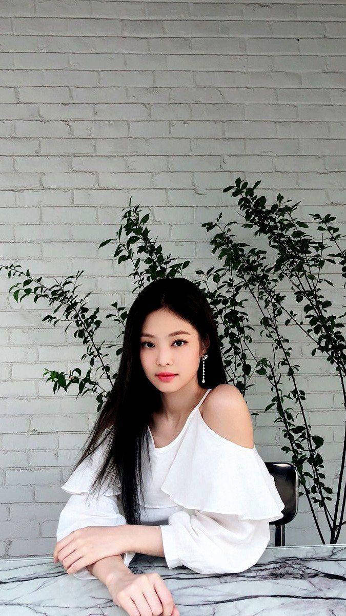 Jennie 桌布 壁紙 — 一位身穿白色露肩上衣的年輕女性坐在大理石桌旁，背景是帶有綠色植物的白色磚牆。