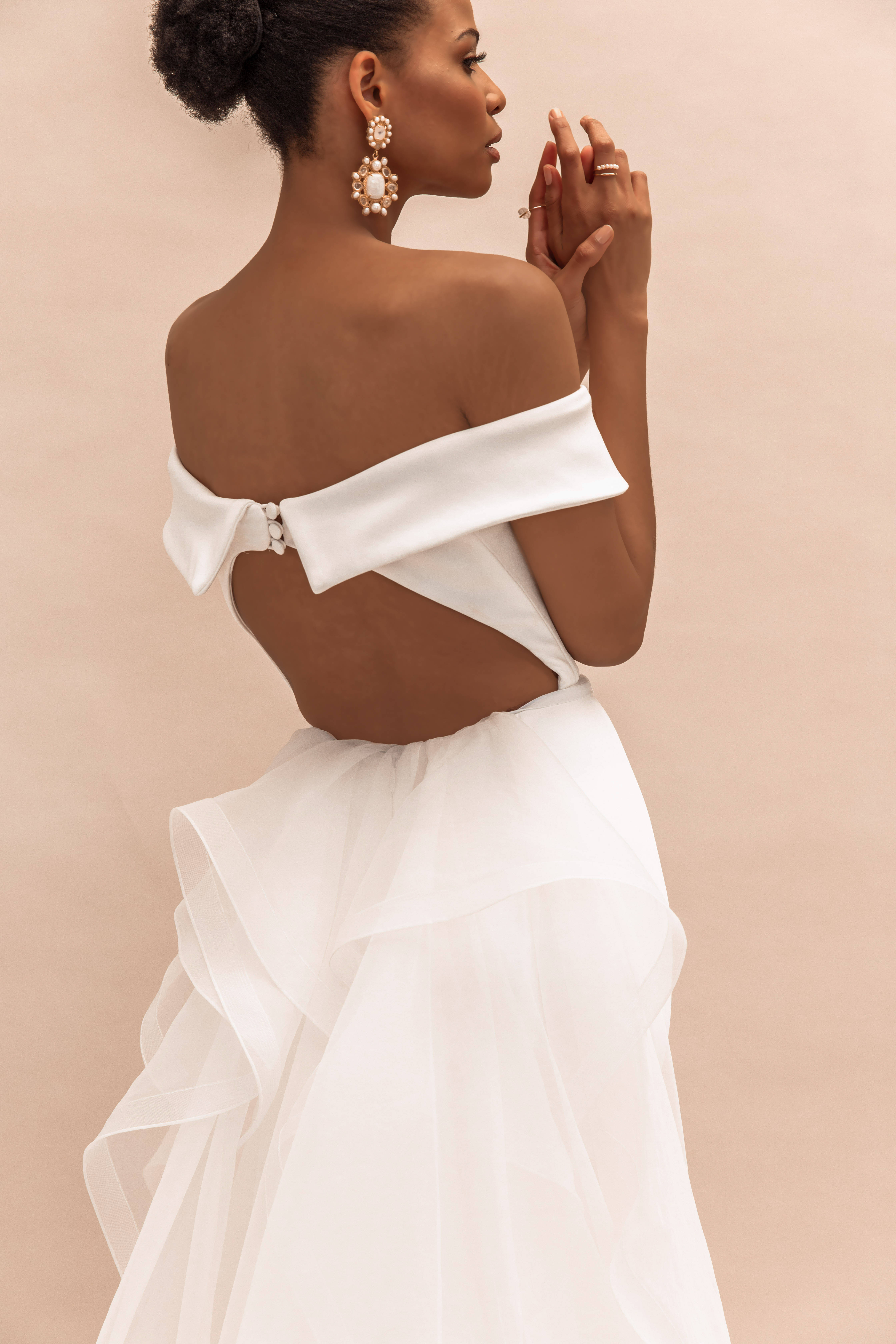 Wedding Aesthetic 桌布 壁紙 — 一位身穿白色露肩禮服、背部鏤空、裙擺荷葉邊的女性，在柔和的米色背景前優雅地擺姿勢。