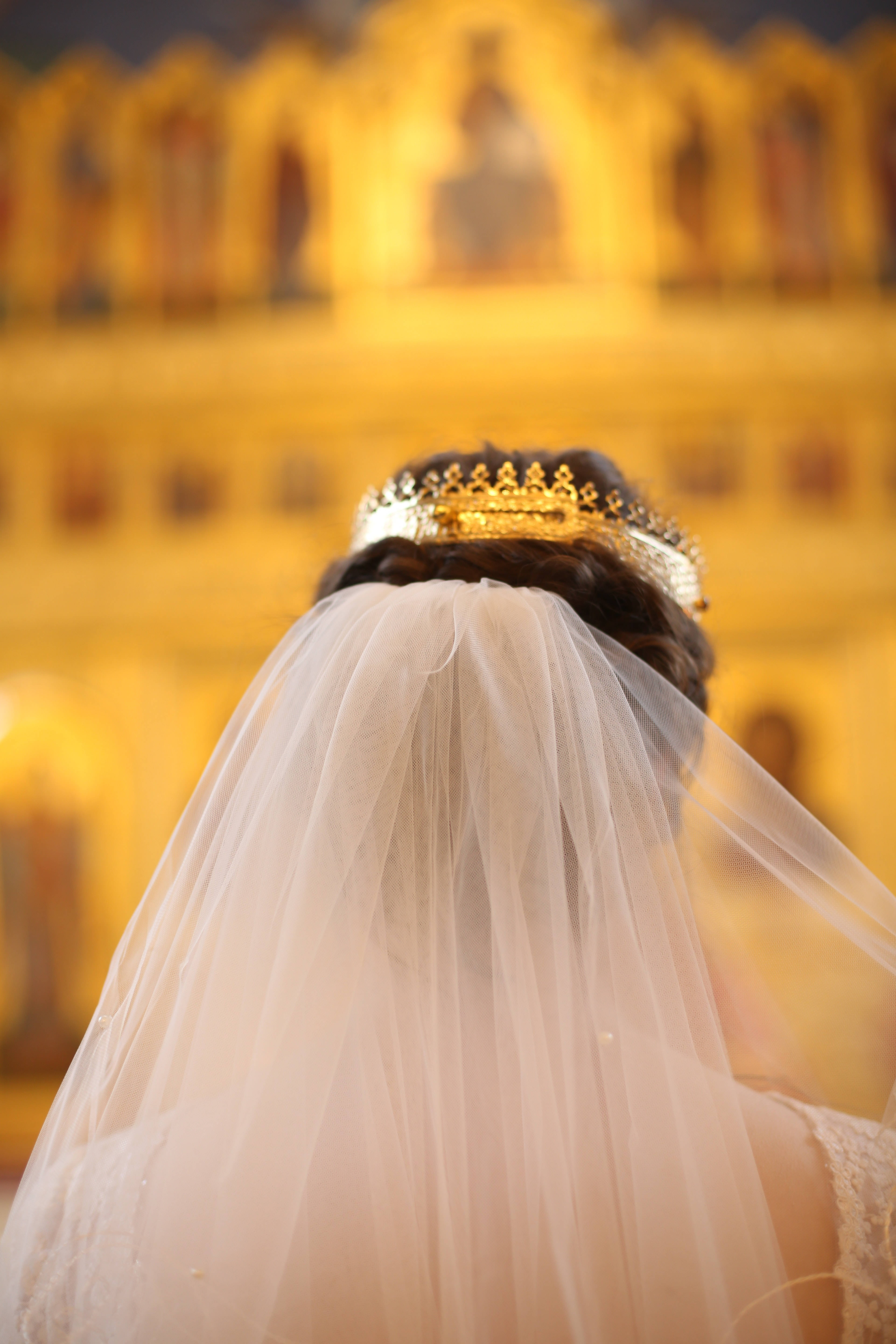 Wedding 桌布 壁紙 — 一位身穿白色頭紗和金色皇冠的新娘背對鏡頭，面向溫暖燈光裝飾華麗的教堂內部。