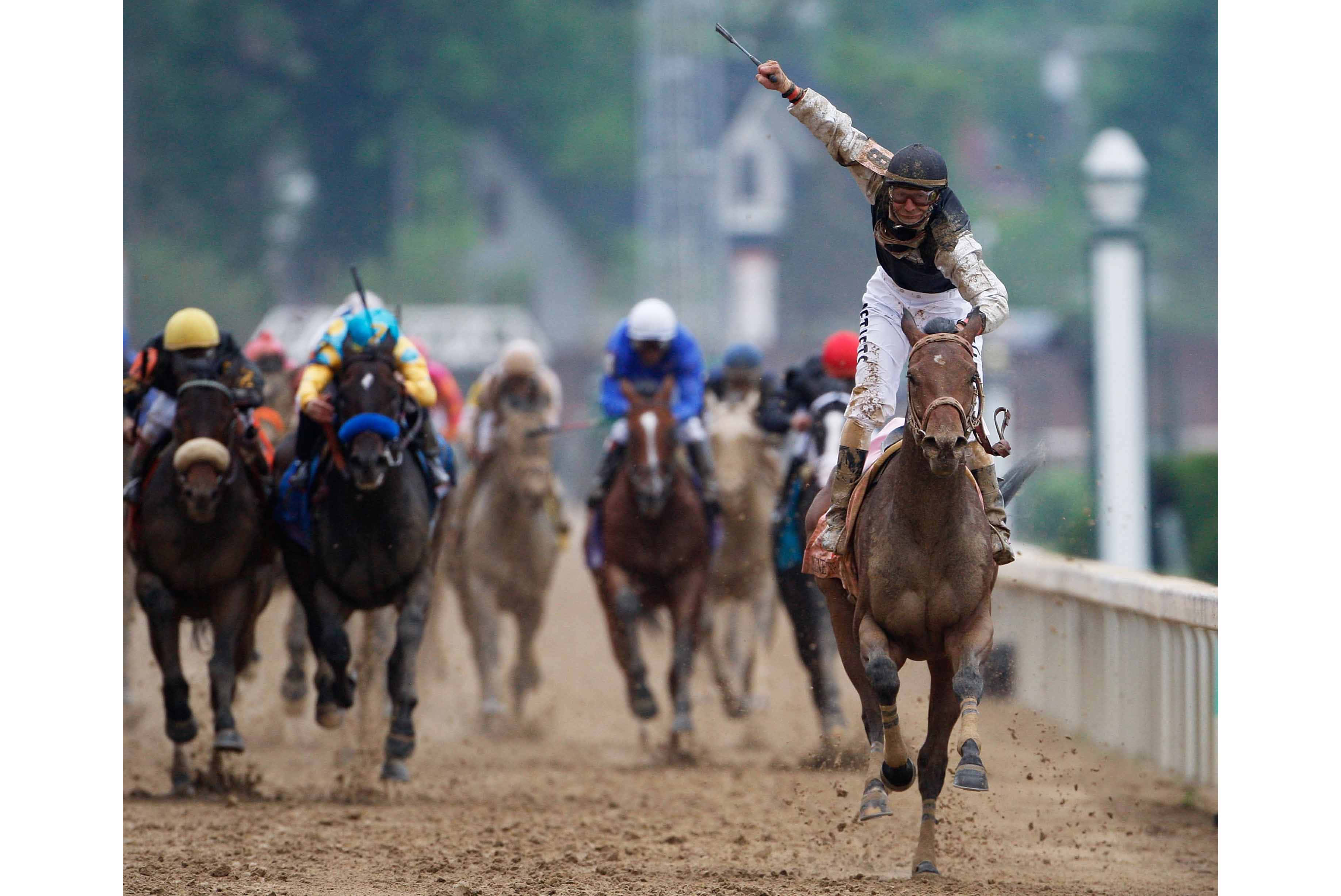 Kentucky Derby 桌布 壁紙 — 一位身穿白色騎師服的騎師在棕色馬上慶祝勝利，其他馬匹在泥濘的賽道上從後追趕。