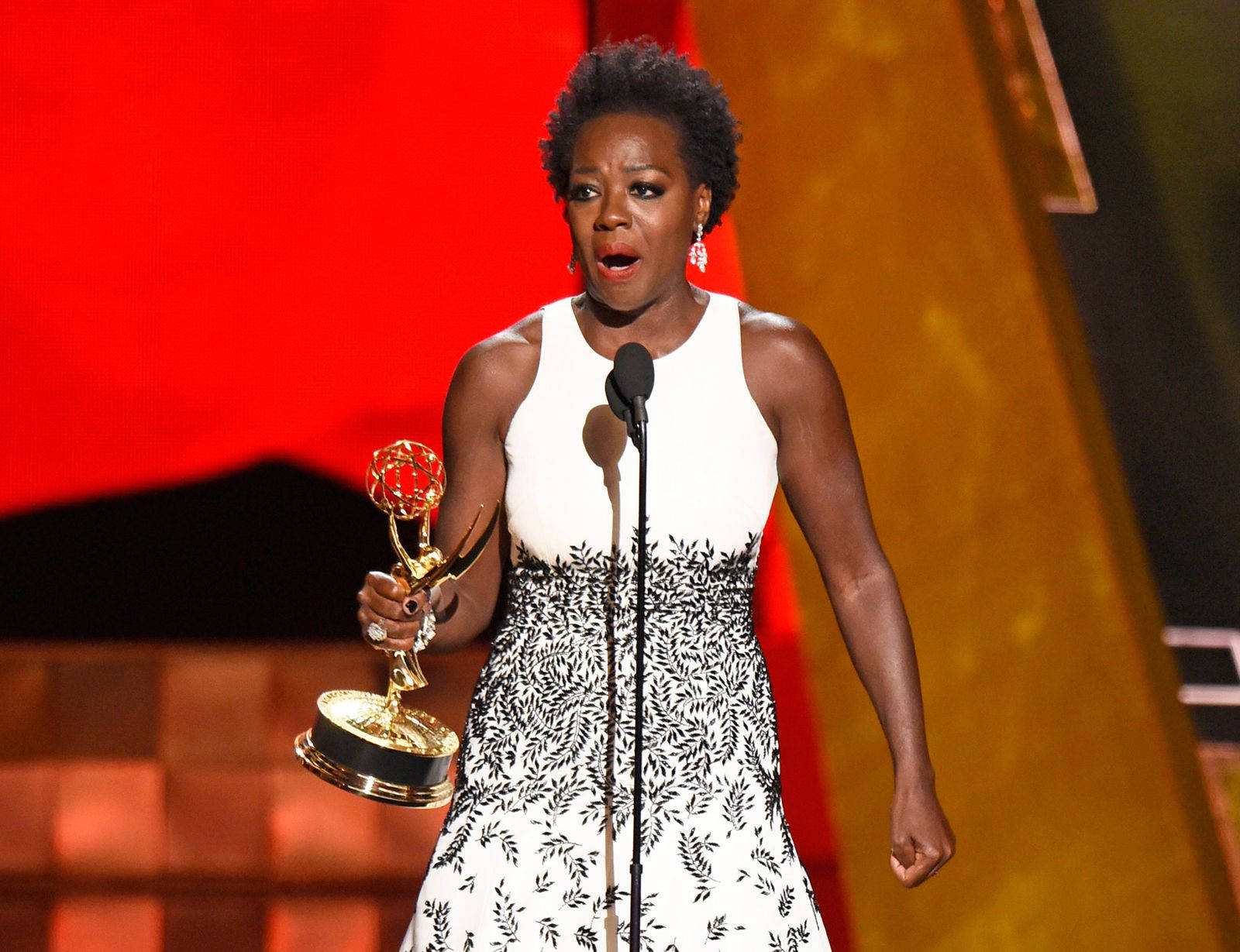 oscars 桌布 壁紙 — 一位身穿白色黑葉圖案連衣裙的女性在紅金色燈光的舞台上手持金色艾美獎獎盃。