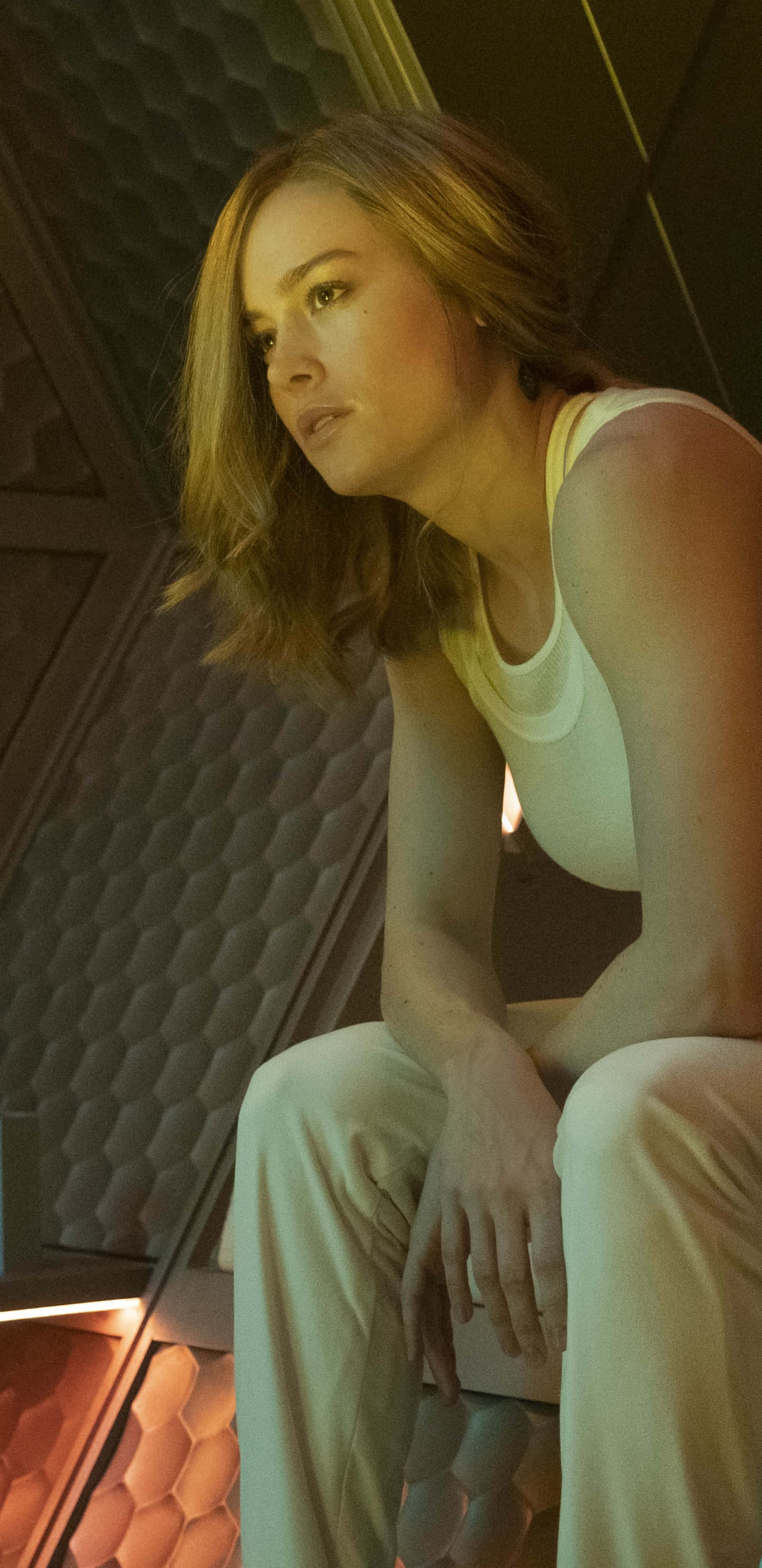 brie larson 桌布 壁紙 — 一位身穿白衣的女性，坐在一個六角形紋理長凳上，沉思於一個昏暗的未來主義室內，帶有溫暖的環境照明。
