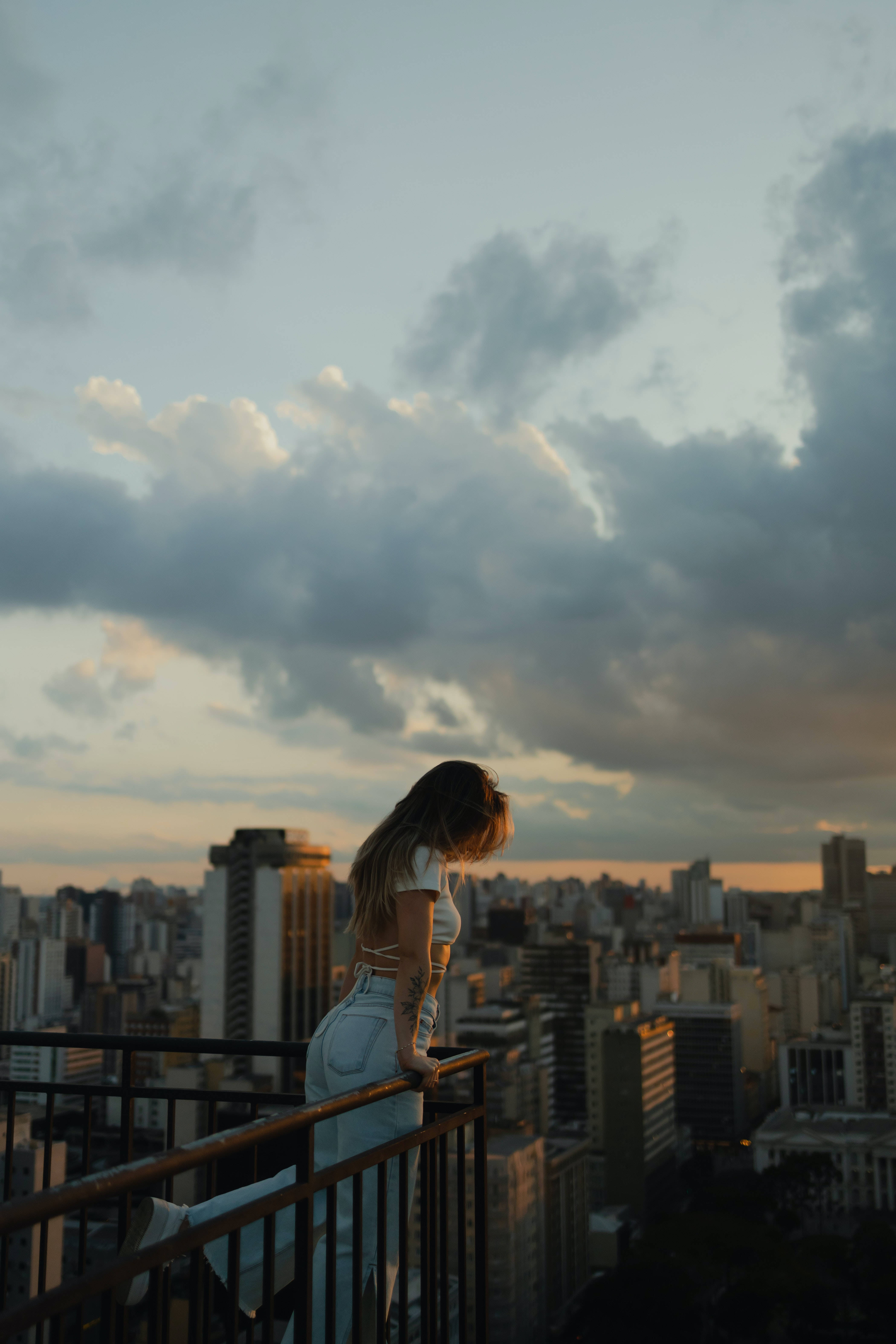 雲朵 桌布 壁紙 — 一位身穿白衣的女性倚在欄杆上，俯瞰夕陽下戲劇性雲彩籠罩的廣闊城市景觀。