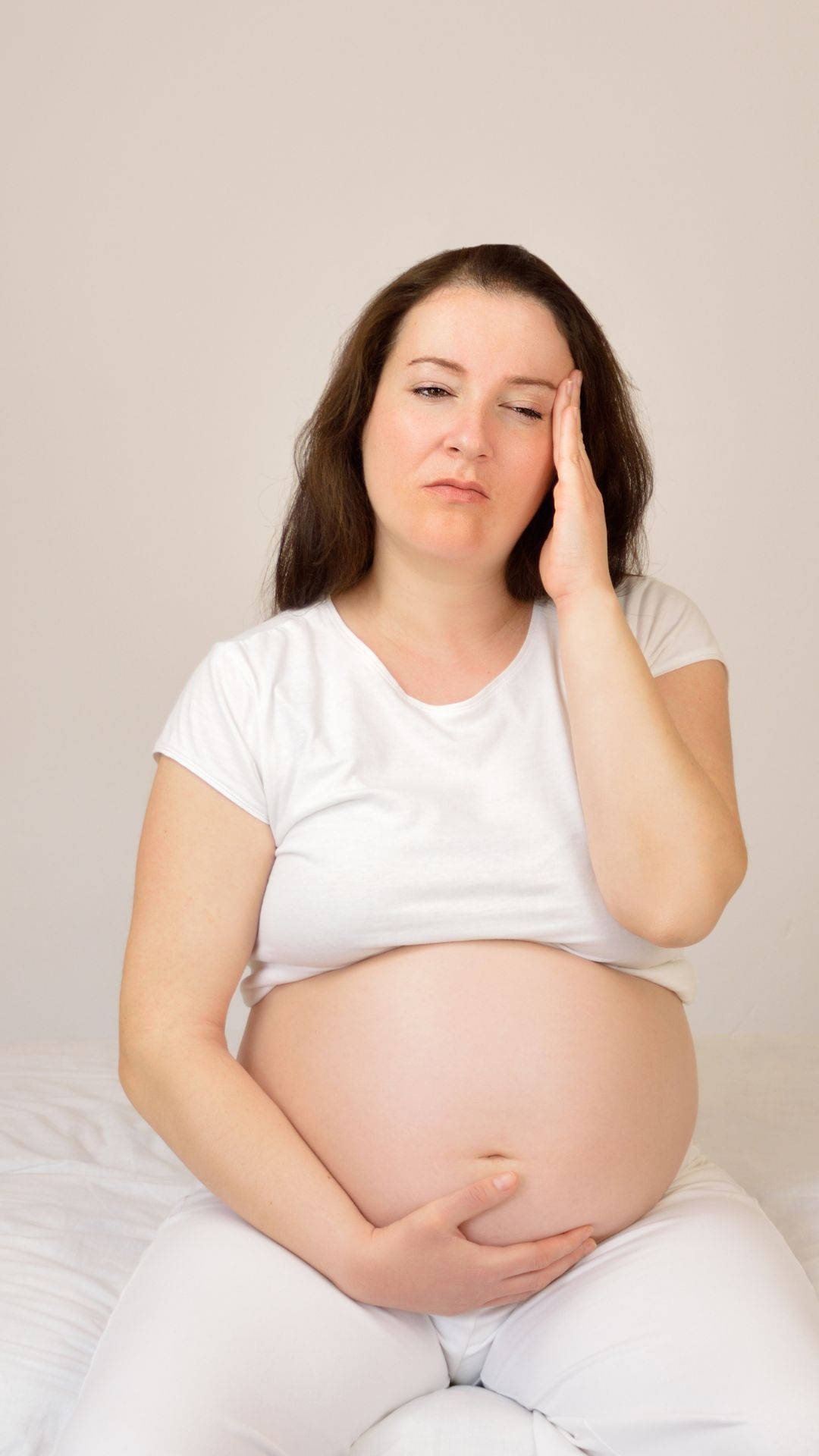 Woman 桌布 壁紙 — 一位身穿白衣的孕婦坐在床上，抱著肚子，額頭靠在手上，露出疲憊的表情。
