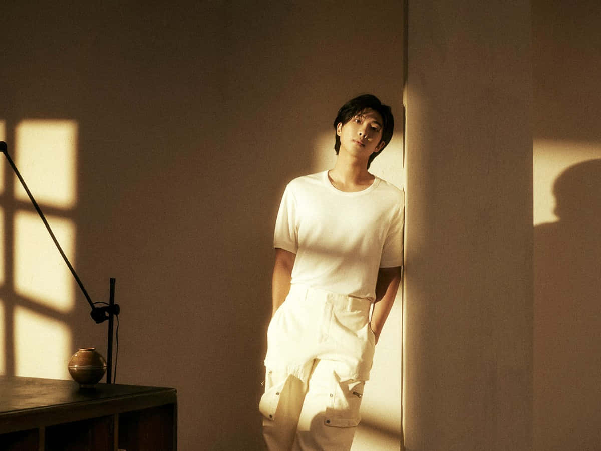 Namjoon 桌布 壁紙 — 一位身穿白衣的年輕男子靠在一面牆上，牆上灑滿了從窗戶射進來的溫暖金色陽光。