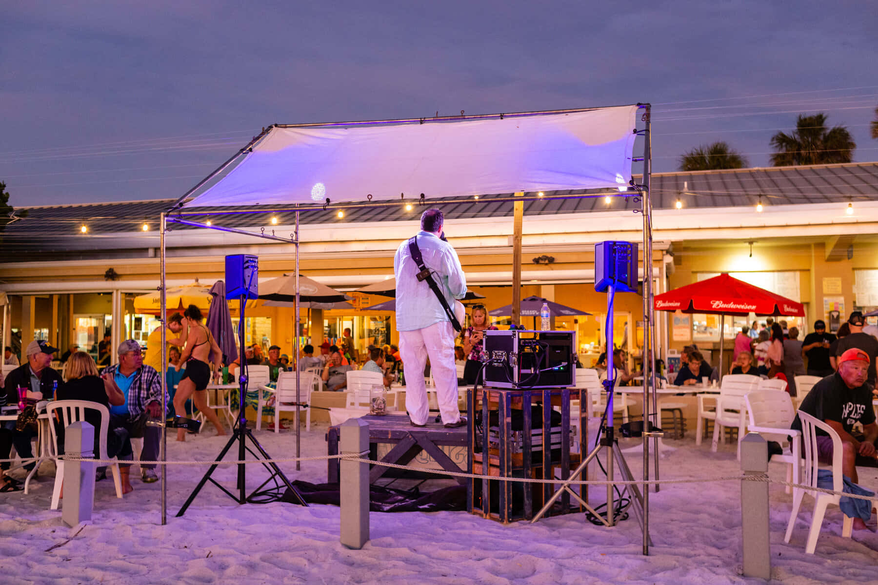 Beach Party 桌布 壁紙 — 一位身穿白衣的音樂家在黃昏的海邊舞台上彈吉他，顧客在戶外餐桌享受飲料和音樂。