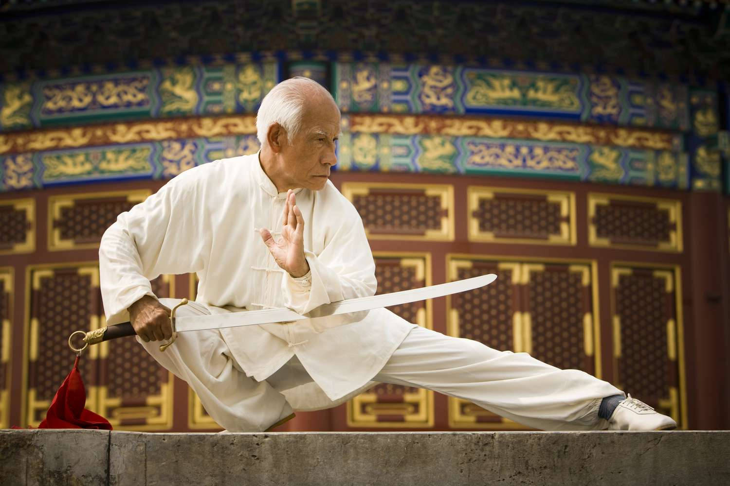 運動 桌布 壁紙 — 一位身穿白袍的老年武術家，在裝飾華麗的傳統中式建築前的石台上，擺出精準的劍式。