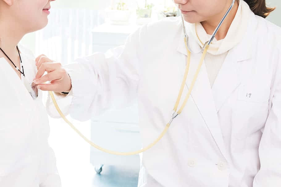 Nurse 桌布 壁紙 — 一位身穿白袍的醫生在明亮的臨床環境中使用聽診器聆聽病人的心跳。