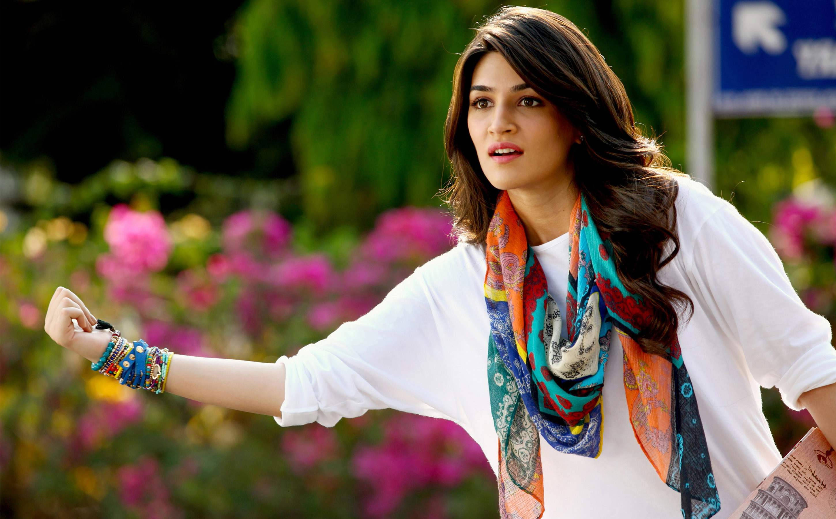 Kriti 桌布 壁紙 — 一位身穿白襯衫和彩色圍巾的年輕女性，站在充滿活力的粉紅色花朵和茂密綠植中做手勢。