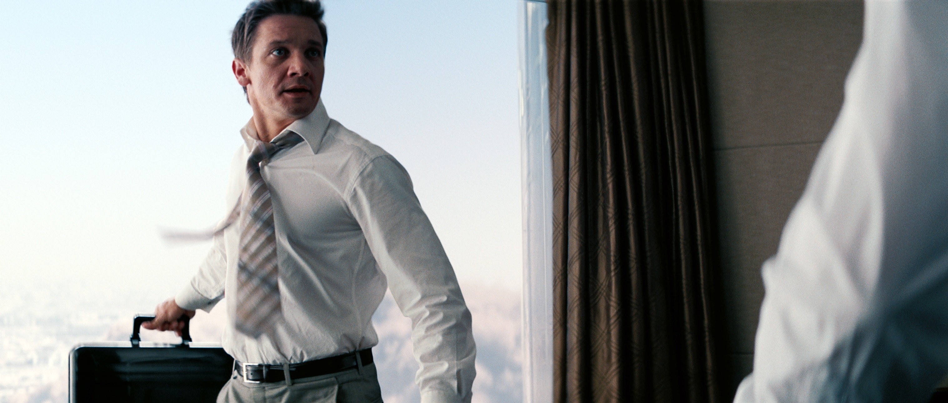 Jeremy Renner 桌布 壁紙 — 一位身穿白襯衫打領帶的商人站在窗邊，手提公事包，領帶隨風飄揚，露出驚訝的表情。
