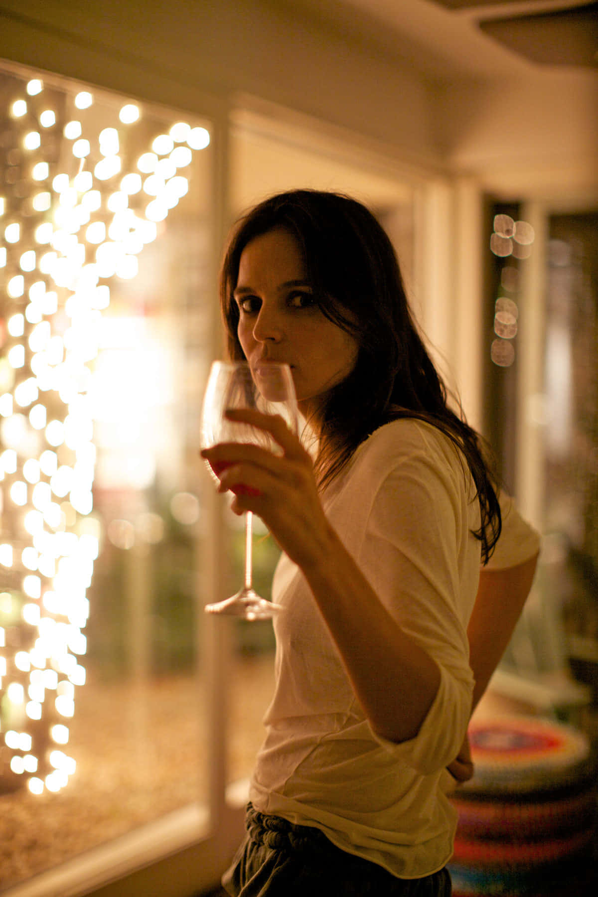Elena Anaya 桌布 壁紙 — 一位身穿白襯衫的女性在室內啜飲葡萄酒，溫暖的串燈在她身後柔和地發光，營造出舒適的夜晚氛圍。
