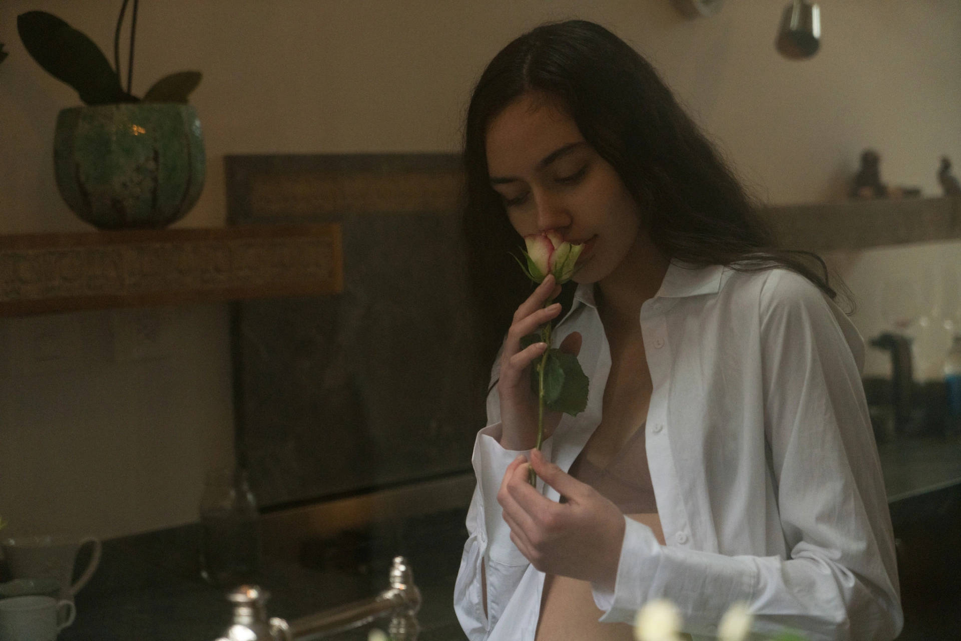 花 桌布 壁紙 — 一位身穿白襯衫的女性在柔和燈光的廚房裡，輕輕聞著玫瑰花香，廚房有木架和盆栽植物。