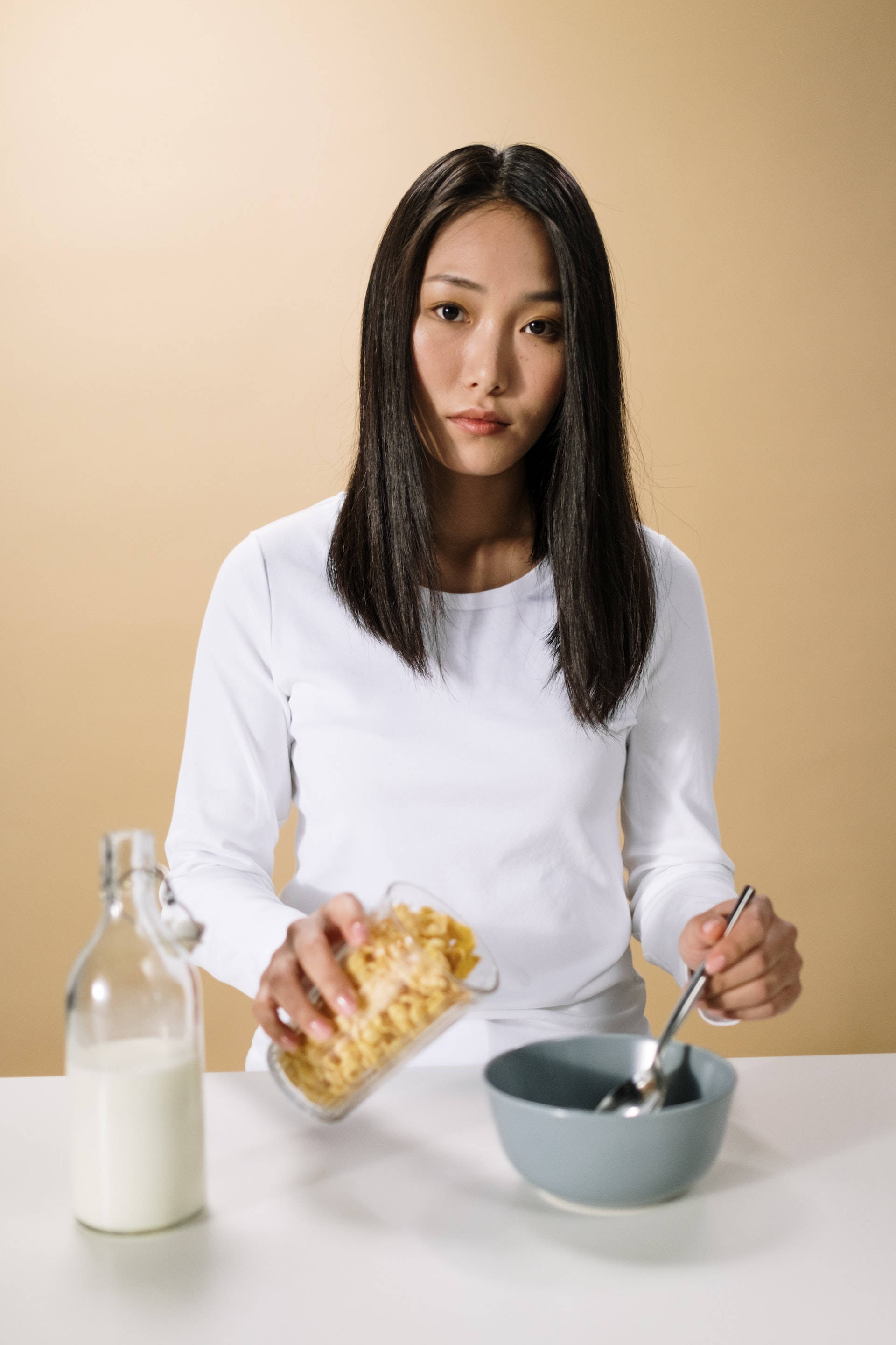 Woman 桌布 壁紙 — 一位身穿白襯衫的女性在米色背景前，將穀物片倒入玻璃牛奶瓶旁邊的碗中。