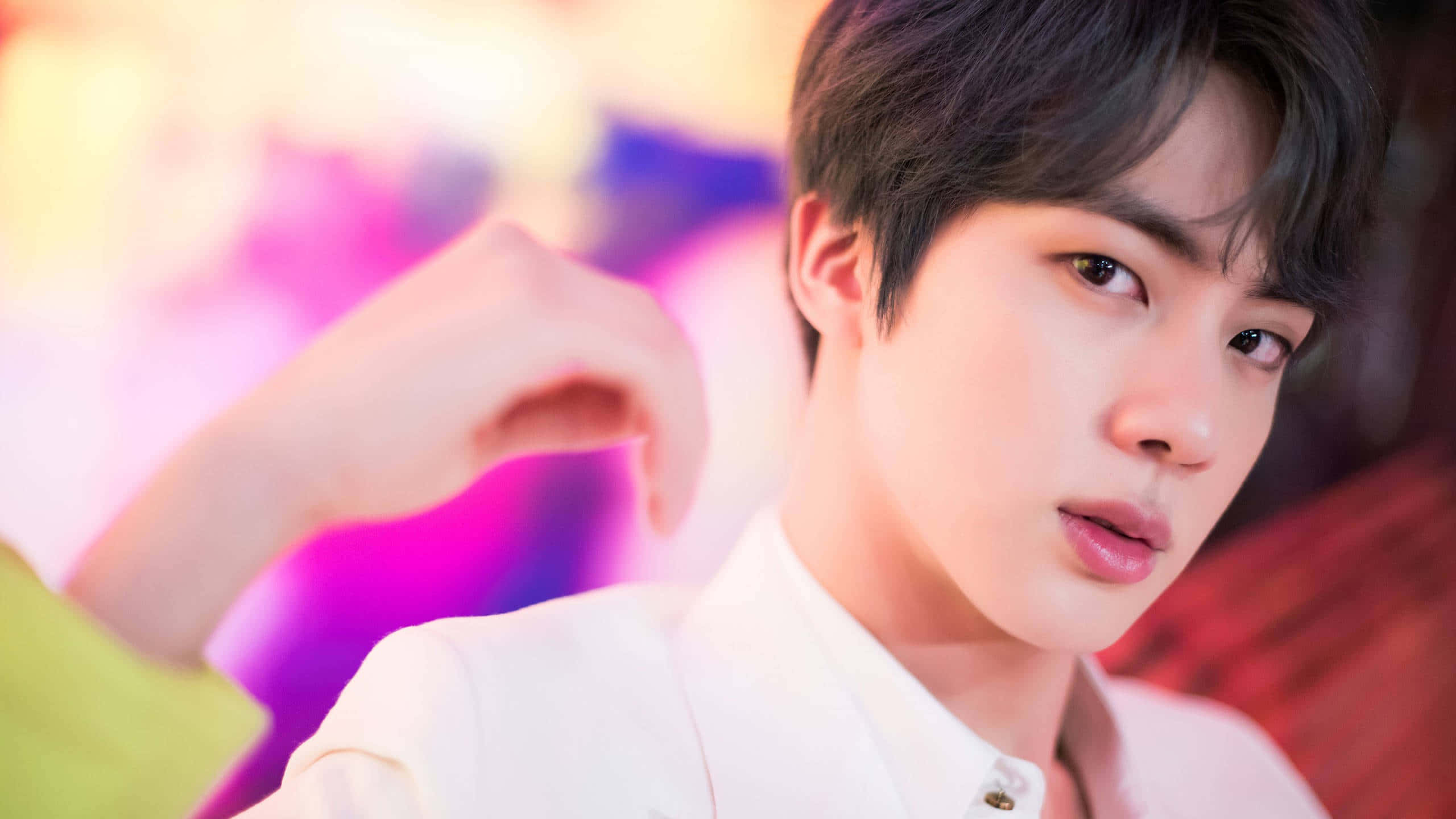 Jin 桌布 壁紙 — 一位身穿白襯衫的年輕男子，在色彩繽紛、充滿活力的模糊背景前，將手靠近臉部擺姿勢。