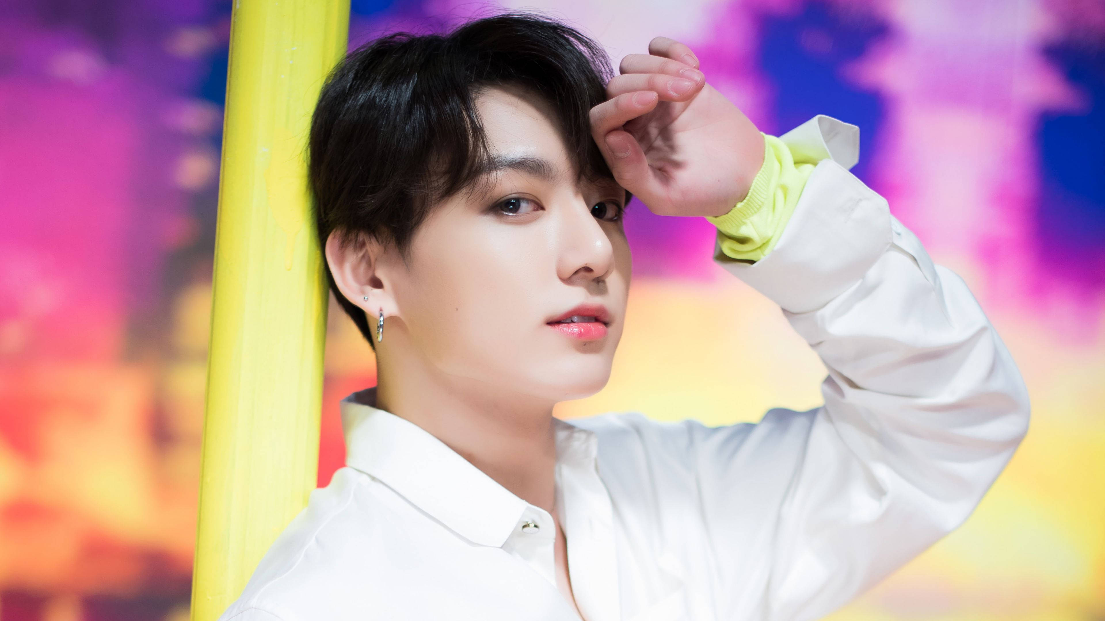 Jungkook 桌布 壁紙 — 一位身穿白襯衫的年輕男子在色彩繽紛的背景前，將手靠近臉部擺姿勢。