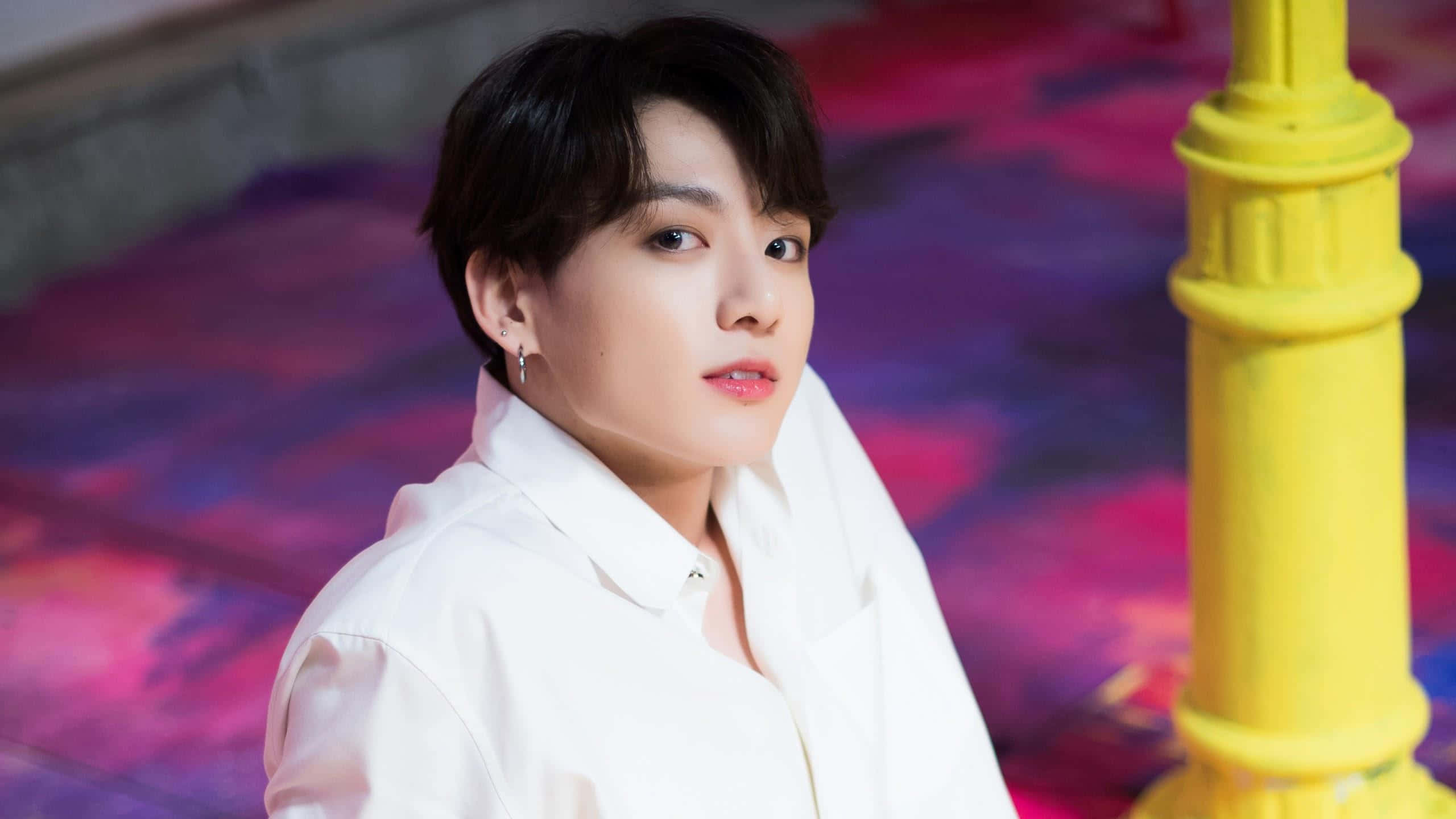 Jungkook 桌布 壁紙 — 一位身穿白襯衫的年輕男子在鮮黃色柱子旁邊，溫柔地凝視著色彩繽紛的紫粉紅色地板。