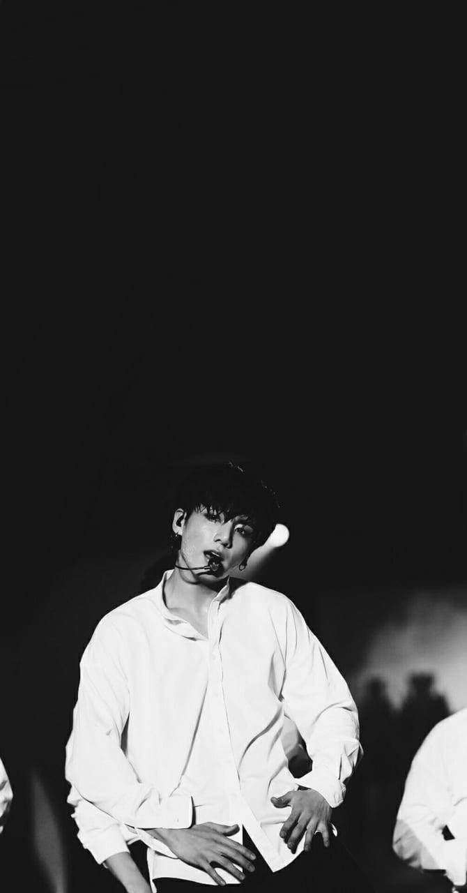 Jungkook Aesthetic 桌布 壁紙 — 一位身穿白襯衫的年輕男子在黑暗的舞台上，在戲劇性的聚光燈下拿著麥克風表演。