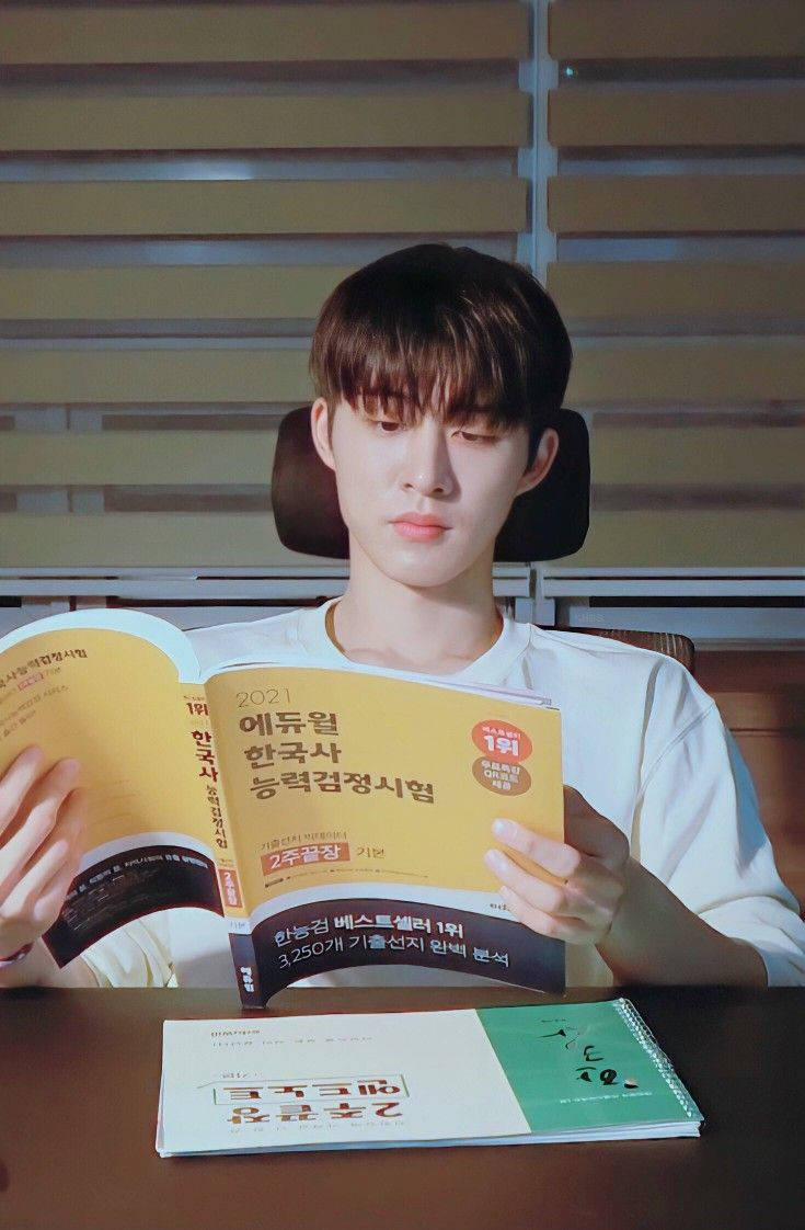 kim hanbin 桌布 壁紙 — 一位身穿白襯衫的年輕男子坐在木桌前，在溫暖的室內燈光下閱讀韓語考試準備書。