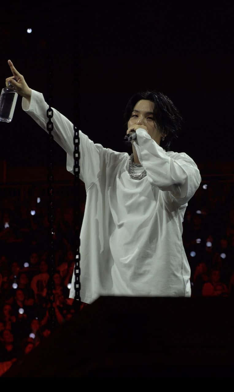Agust D 桌布 壁紙 — 一位身穿白襯衫的表演者，在舞台燈光下拿著酒杯，指向上方，對著麥克風唱歌。