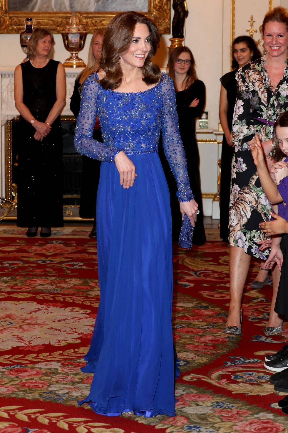 Kate Middleton 桌布 壁紙 — 一位身穿皇家藍色珠飾長裙的女性，在華麗的地毯和鍍金裝飾的豪華宮殿大廳中優雅地微笑。