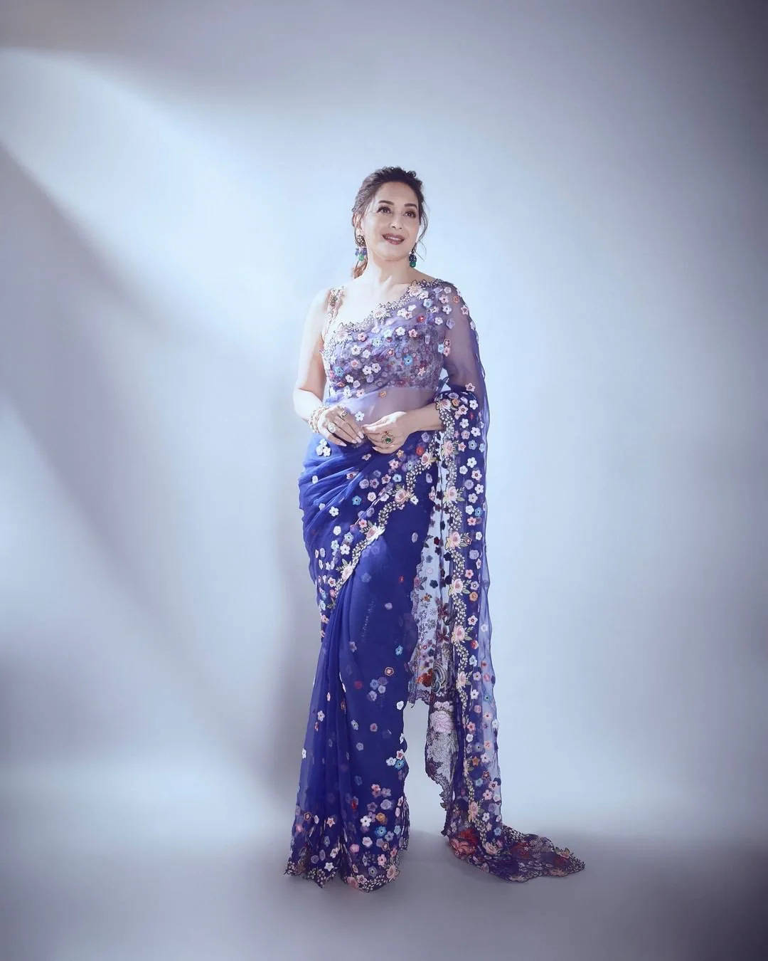 Madhuri Dixit 桌布 壁紙 — 一位身穿皇家藍花卉紗麗、帶有透明裝飾的女性，在柔和的灰色攝影棚背景下優雅地站立。