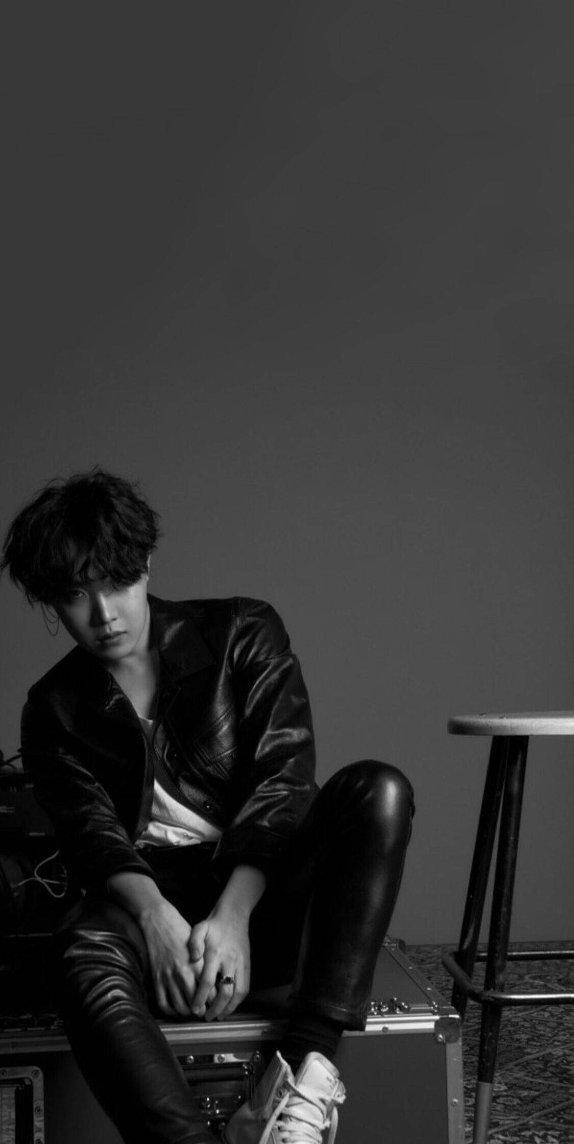 j-hope 桌布 壁紙 — 一位身穿皮夾克的年輕男子，在單色工作室拍攝中，沉思地坐在一個齒輪箱上，背景是純灰色的牆壁。