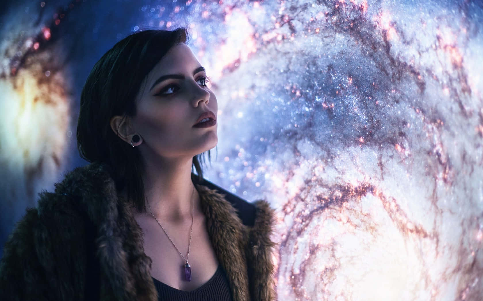 Celestial 桌布 壁紙 — 一位身穿皮草大衣的女性仰望一個旋轉的宇宙星雲，散發著藍色和粉紅色的光芒。