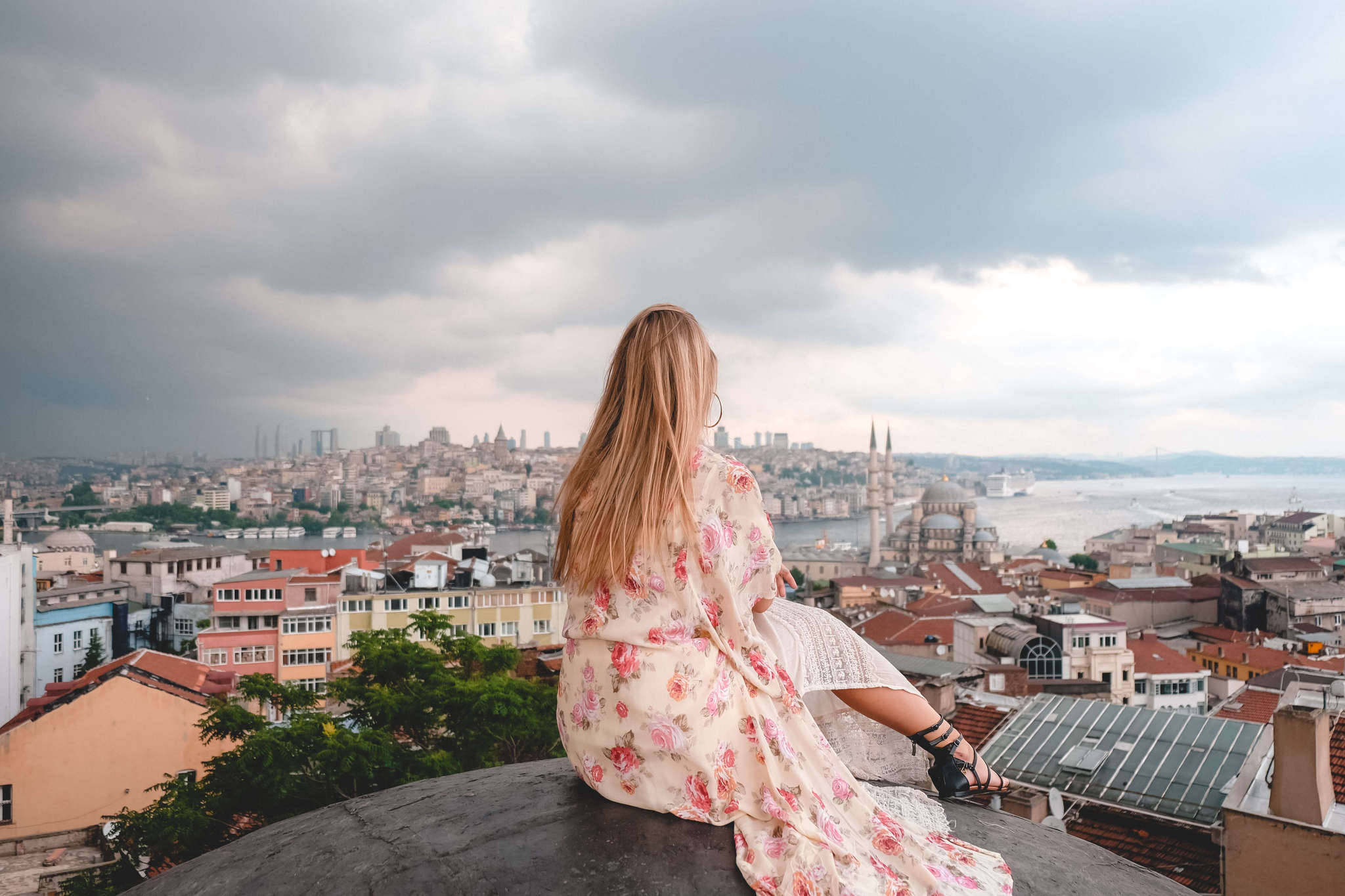 Istanbul 桌布 壁紙 — 一位身穿碎花裙的女士坐在屋頂上，俯瞰多雲天空下的廣闊城市景觀。