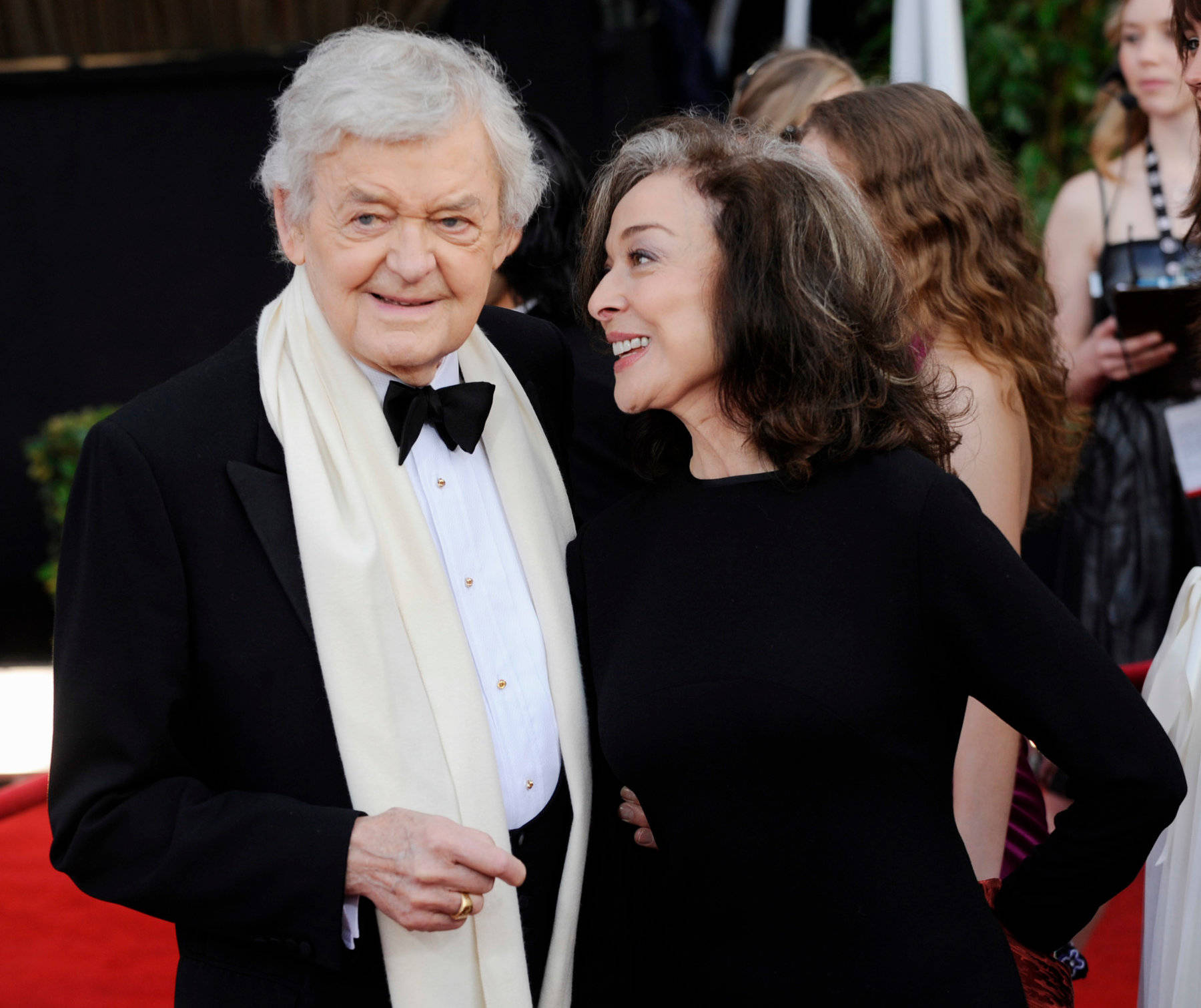 Celebrity 桌布 壁紙 — 一位身穿禮服、戴著白色圍巾的長者男士，在紅地毯上與一位身穿黑色連衣裙的女士旁邊溫暖地微笑。