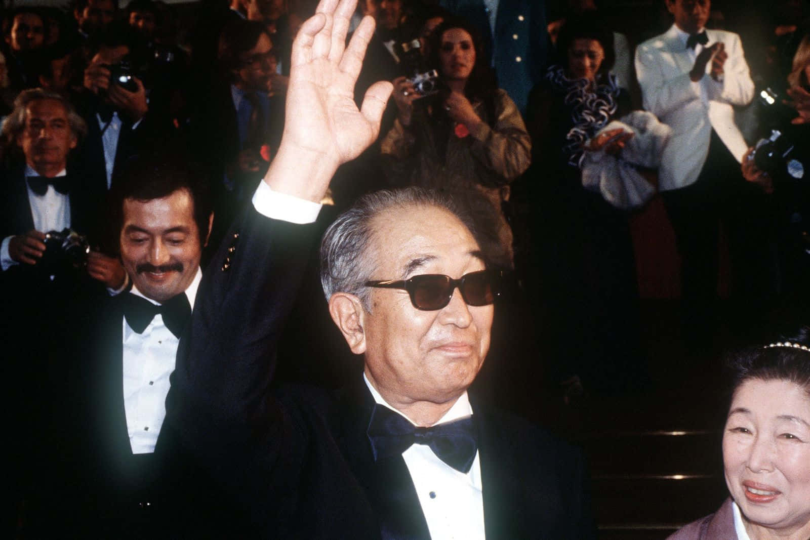 Akira Kurosawa 桌布 壁紙 — 一位身穿禮服和太陽眼鏡的長者，在一個華麗的活動中向一群攝影師和旁觀者揮手。