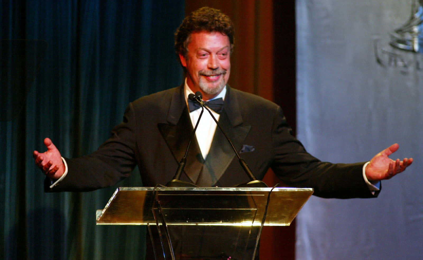 Tim Curry 桌布 壁紙 — 一位身穿禮服的微笑男士在頒獎典禮上張開雙臂站在講台上。
