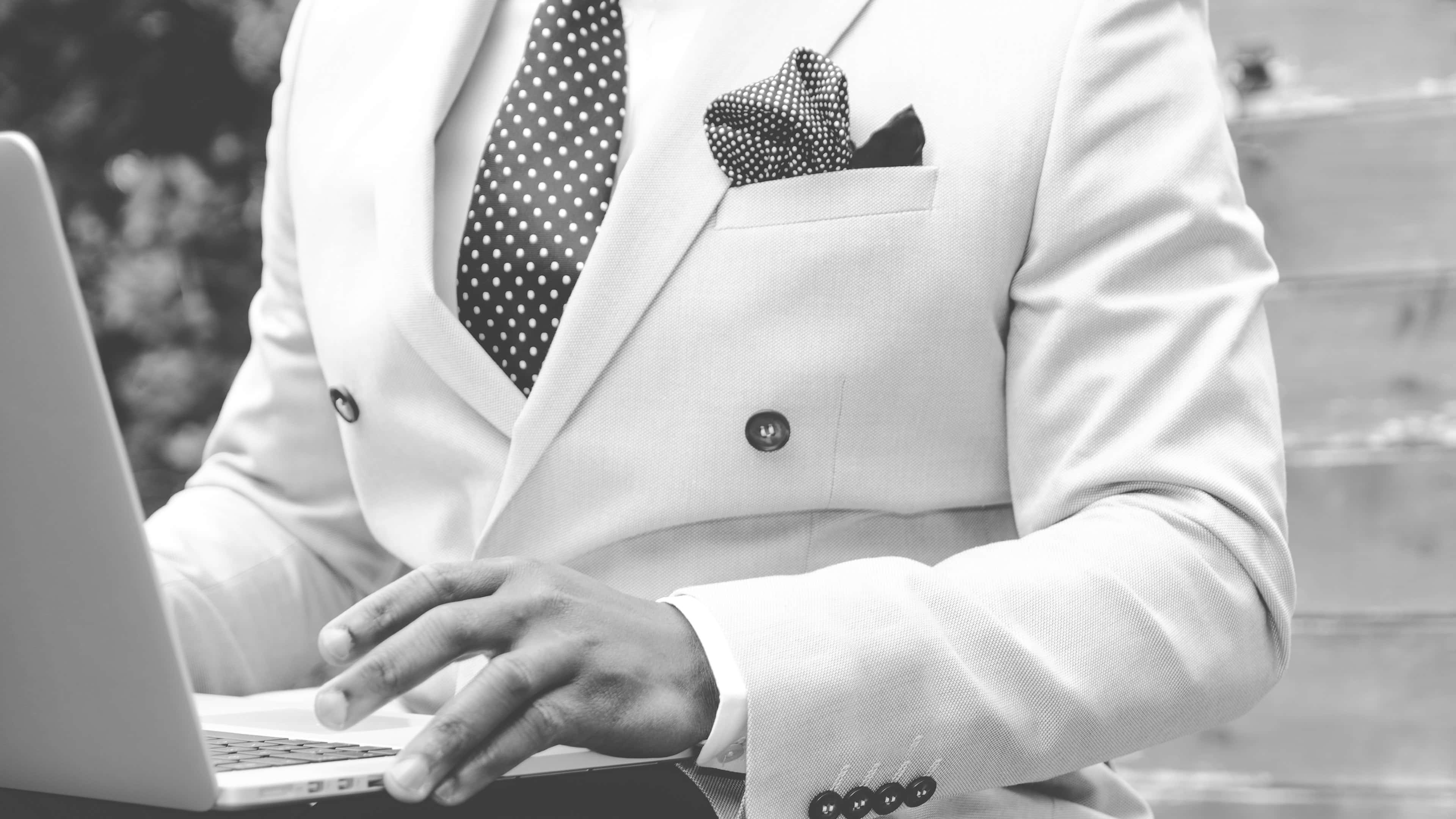 style 桌布 壁紙 — 一位身穿筆挺白色西裝、打著波點領帶的男士，在戶外單色調環境下使用筆記型電腦打字。