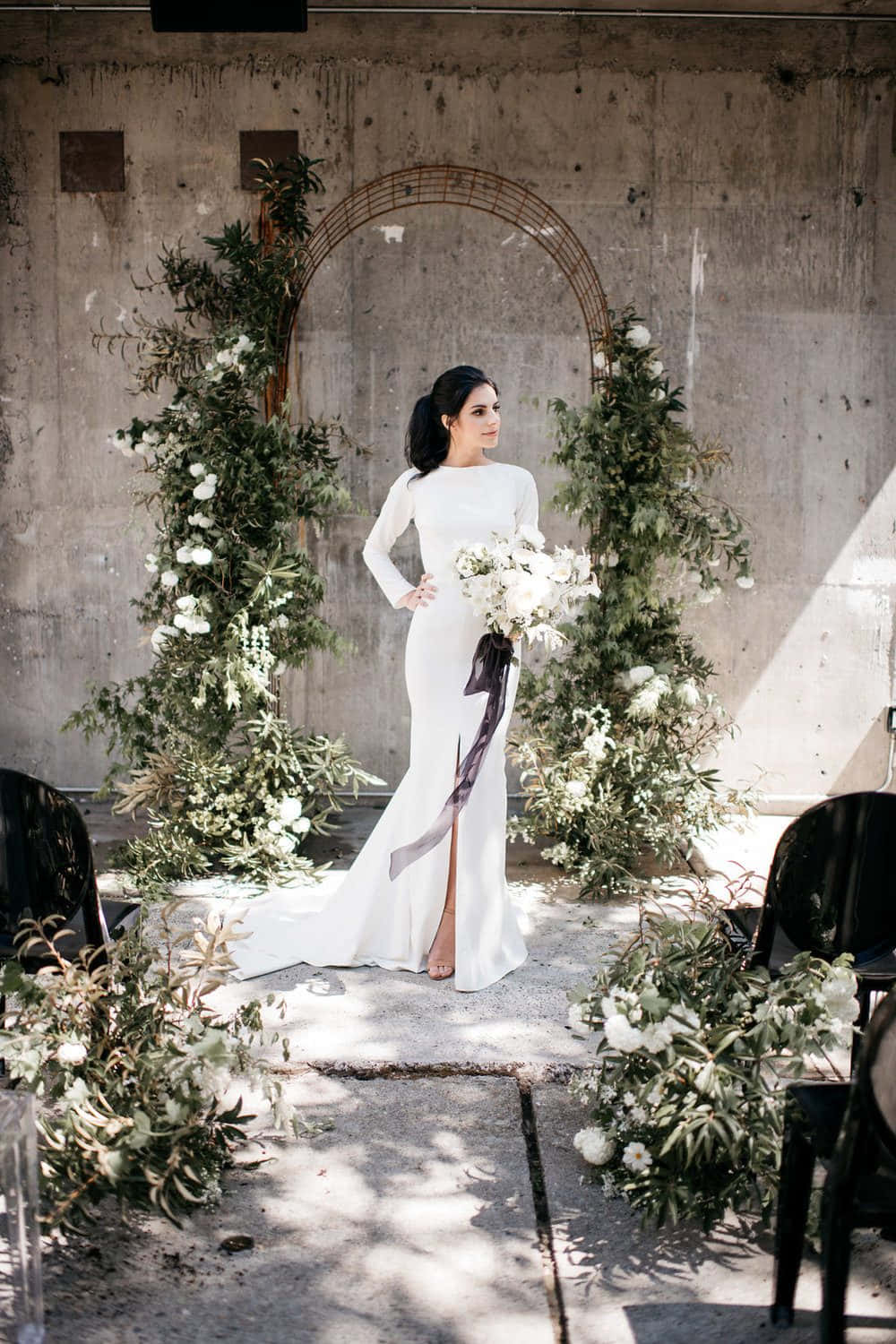 Black And White Wedding 桌布 壁紙 — 一位身穿簡潔白色婚紗的新娘，自信地站在質樸的花拱門下，背景是風化的混凝土牆。