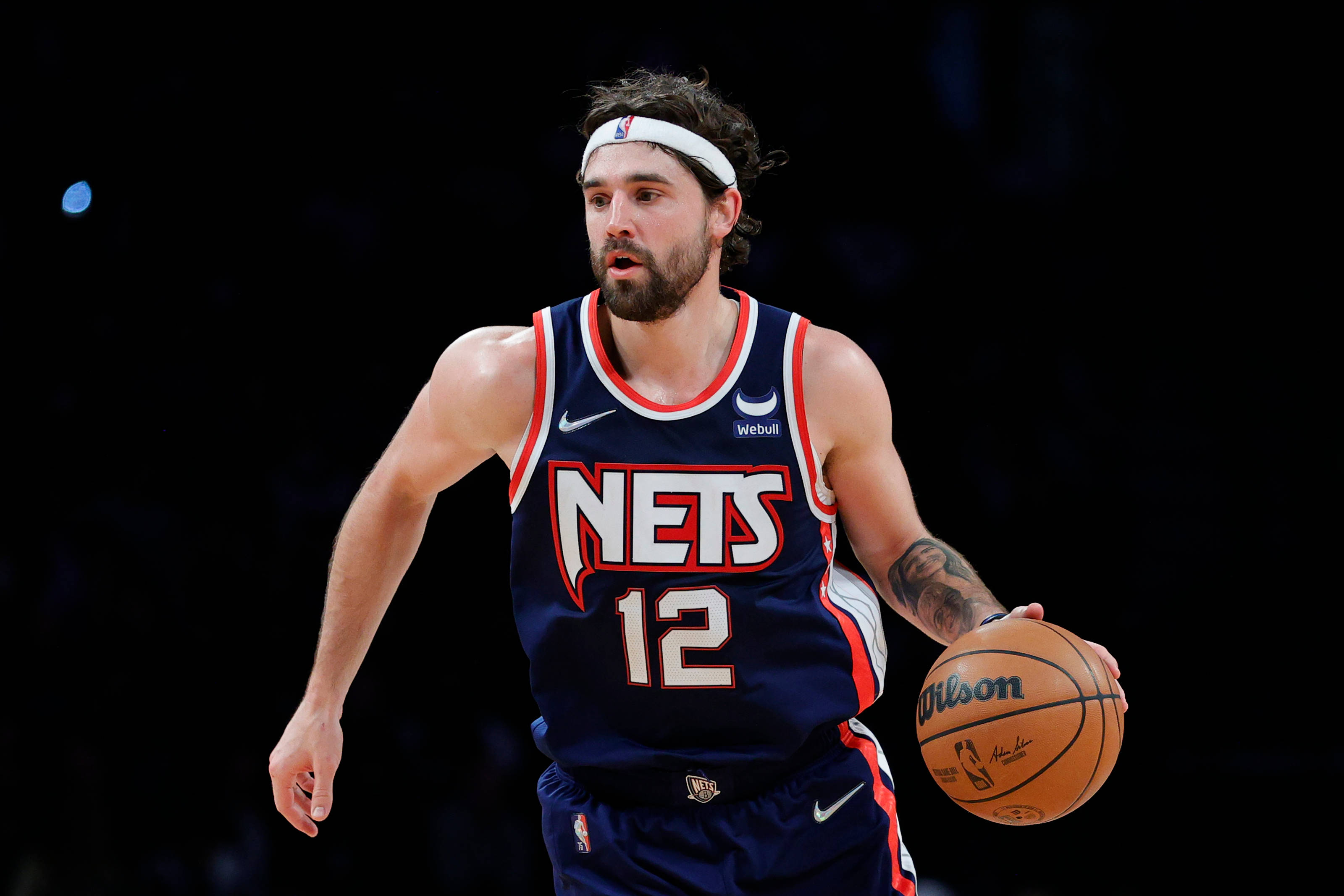 joe harris 桌布 壁紙 — 一位身穿籃網隊球衣的籃球運動員在黑暗背景下，於場館燈光下運著威爾遜籃球。