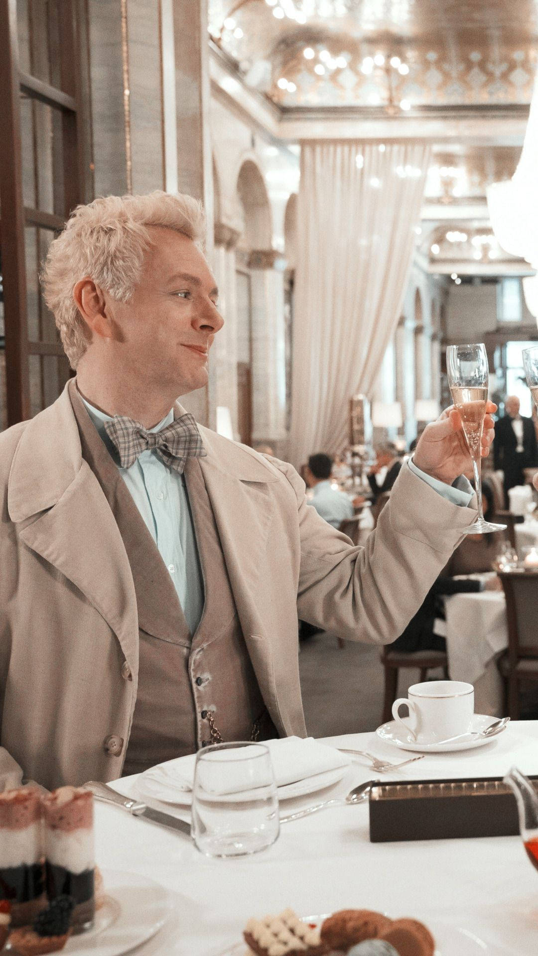 Good Omens 桌布 壁紙 — 一位身穿米色外套和領結的男子，在一間裝飾華麗、燈光溫暖、有客人用餐的宏偉餐廳中舉起香檳杯。