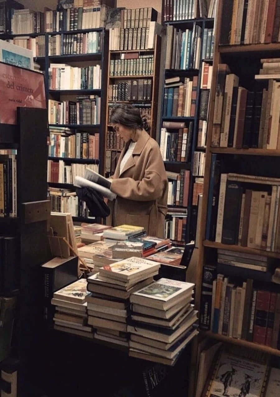 Library 桌布 壁紙 — 一位身穿米色外套的女性在舒適、燈光昏暗的書店中，在堆滿復古書籍的高聳書架間閱讀一本書。