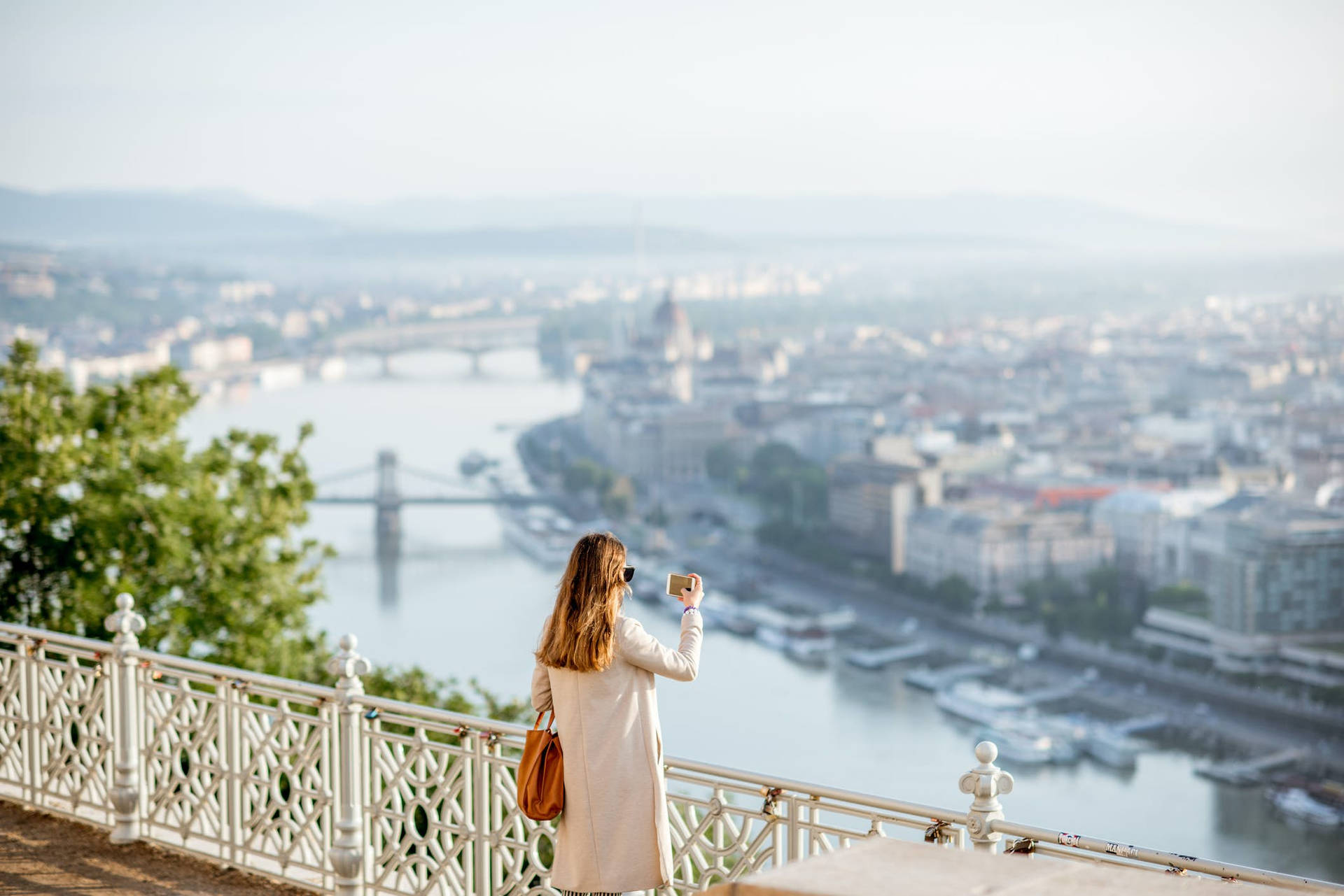 Hungary 桌布 壁紙 — 一位身穿米色外套的女性站在白色欄杆上，俯瞰下方有河流和橋樑的朦朧城市景觀。