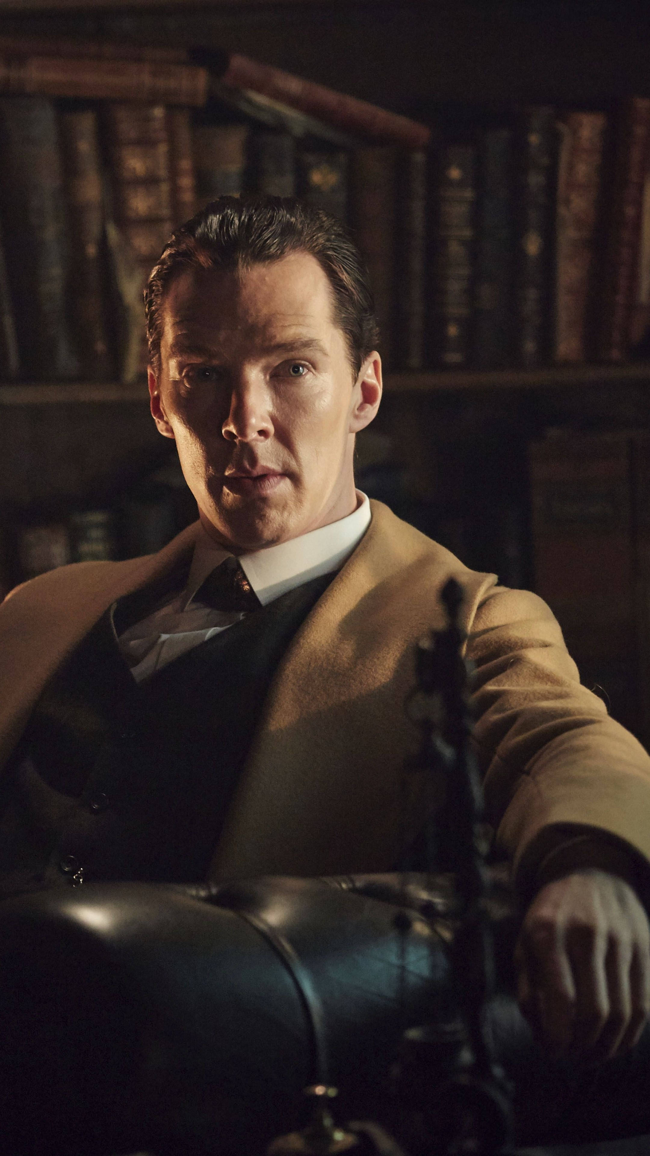 benedict 桌布 壁紙 — 一位身穿米色外套的男子坐在昏暗書房的皮椅上，身後有書架。