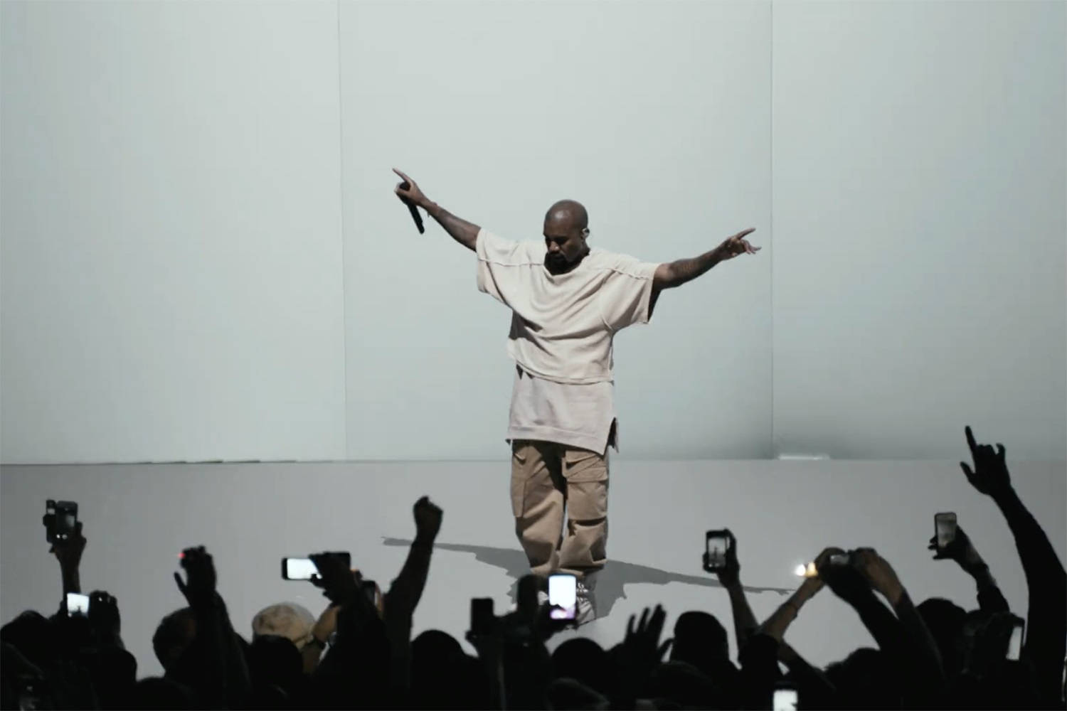 Kanye West Donda 桌布 壁紙 — 一位身穿米色服裝的表演者站在舞台中央，雙臂伸展，而下面的觀眾舉起手機捕捉這一刻。
