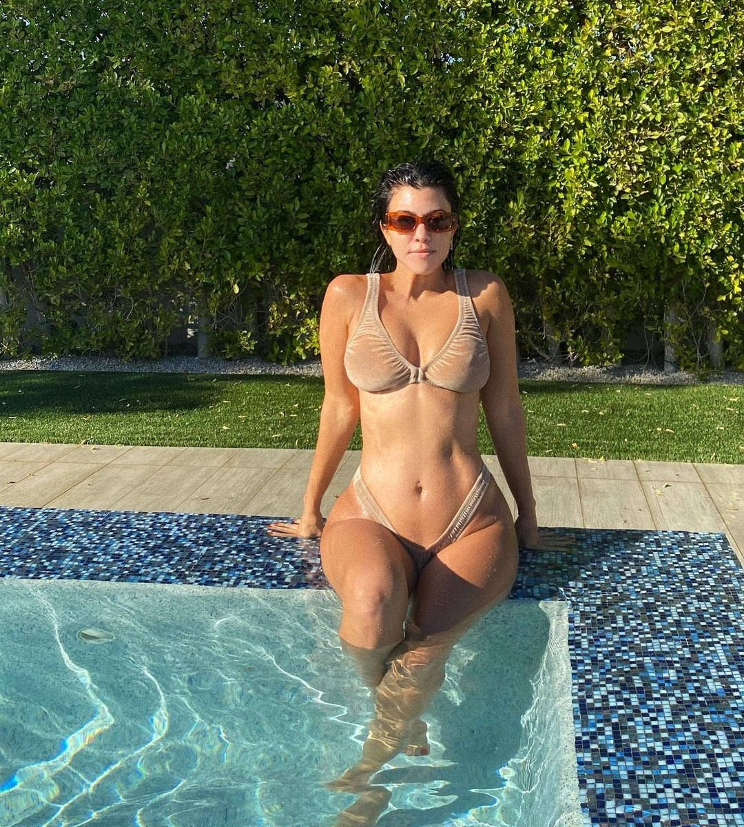 Kourtney 桌布 壁紙 — 一位身穿米色比基尼和太陽眼鏡的女性坐在藍色瓷磚泳池邊，身後是茂密的綠色樹籬。
