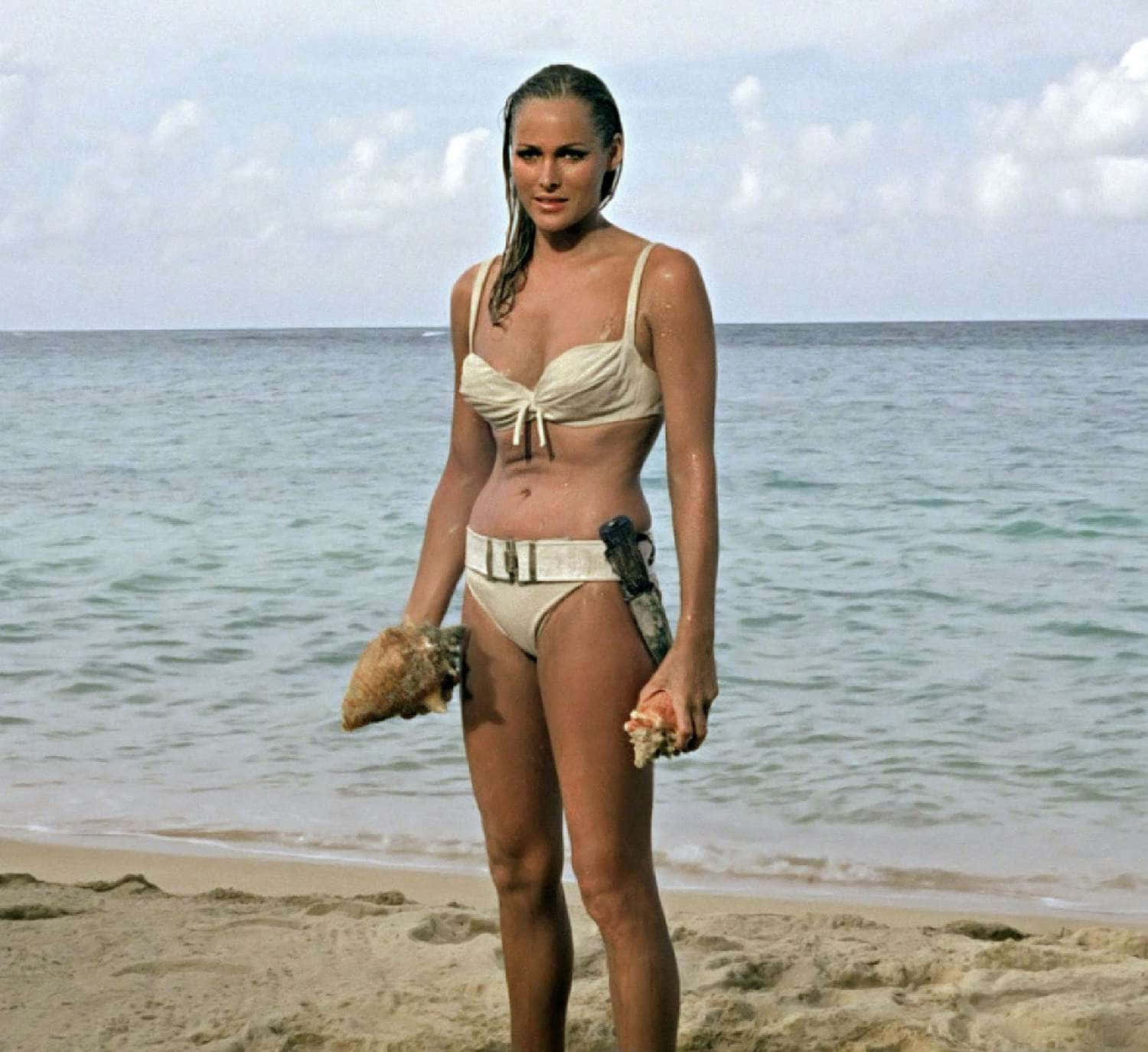 Swimsuit 桌布 壁紙 — 一位身穿米色比基尼的女性站在沙灘上，手持貝殼，身後是海浪。