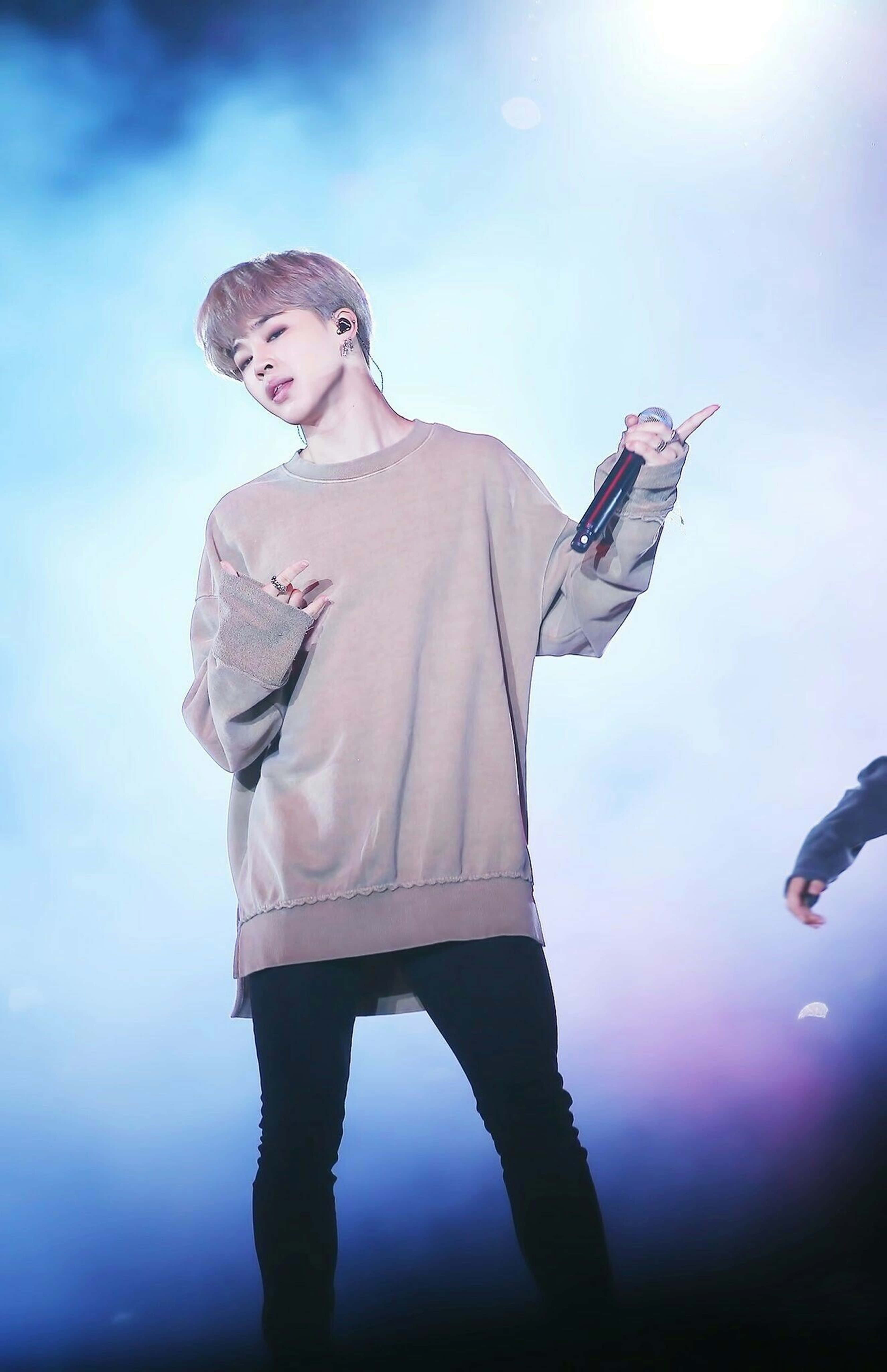 Bts Jimin 桌布 壁紙 — 一位身穿米色毛衣和黑色褲子的表演者在明亮的藍色舞台燈光及煙霧效果下拿著麥克風。