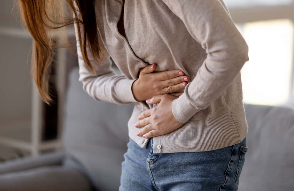生病 桌布 壁紙 — 一位身穿米色毛衣的女性在柔和光線的室內空間中站立，明顯不適地捂著腹部。