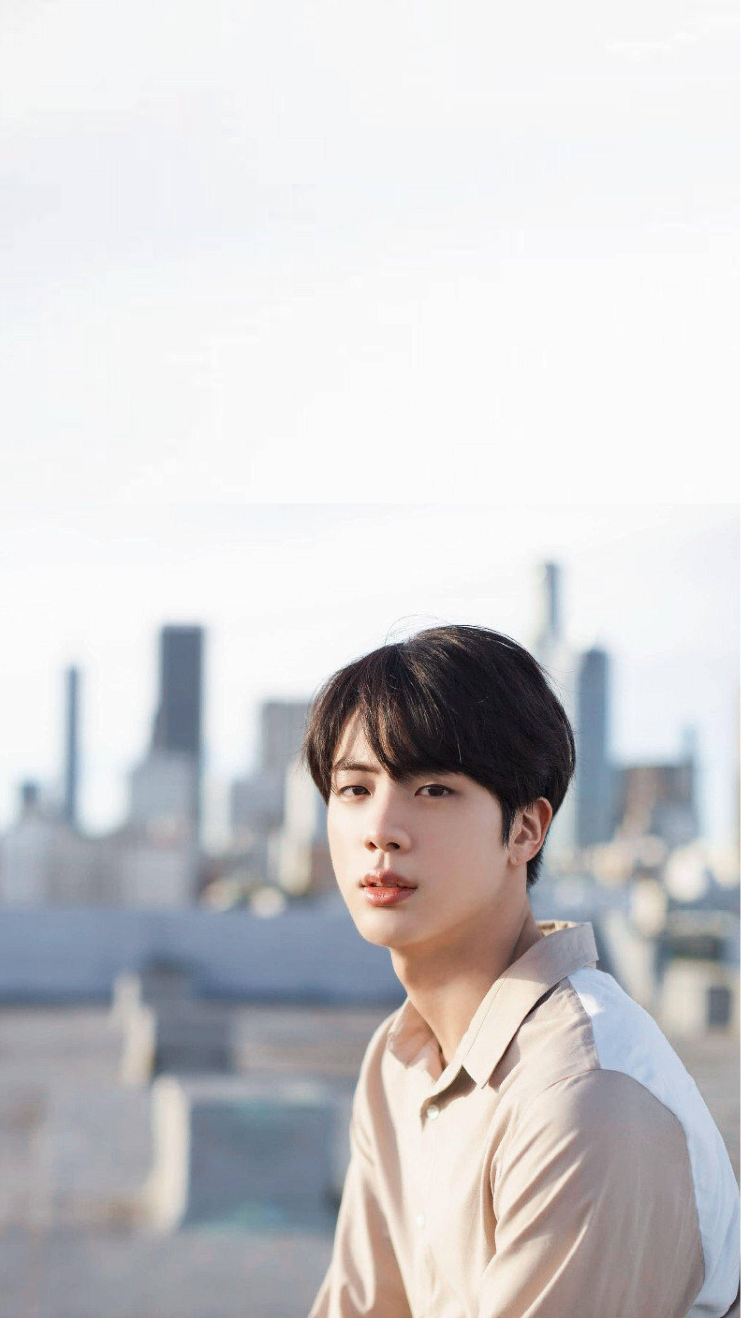 Jin 桌布 壁紙 — 一位身穿米色襯衫的年輕男子在明亮的日光下，若有所思地凝視著朦朧的城市天際線。