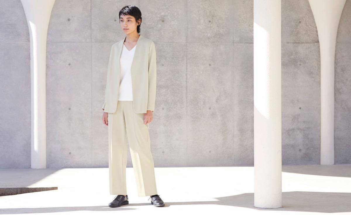 Uniqlo 桌布 壁紙 — 一位身穿米色西裝的女性在陽光下，於白色柱子之間，背靠混凝土牆，靜靜站立。