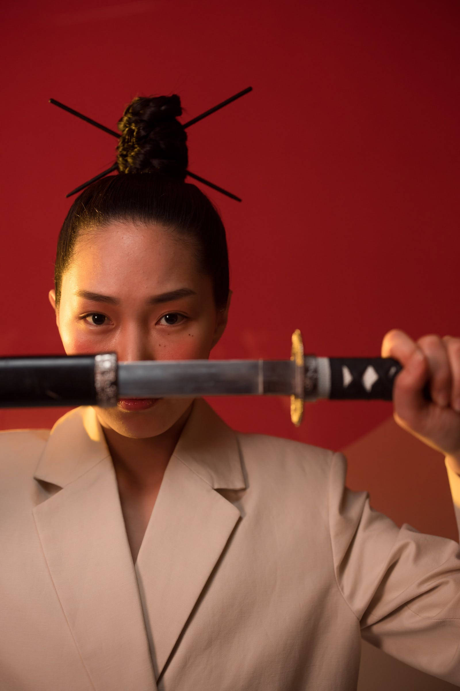 桌布 壁紙 — 一位身穿米色西裝的女性水平握著一把武士刀，她強烈的目光與觀眾對視，背景是鮮明的紅色。
